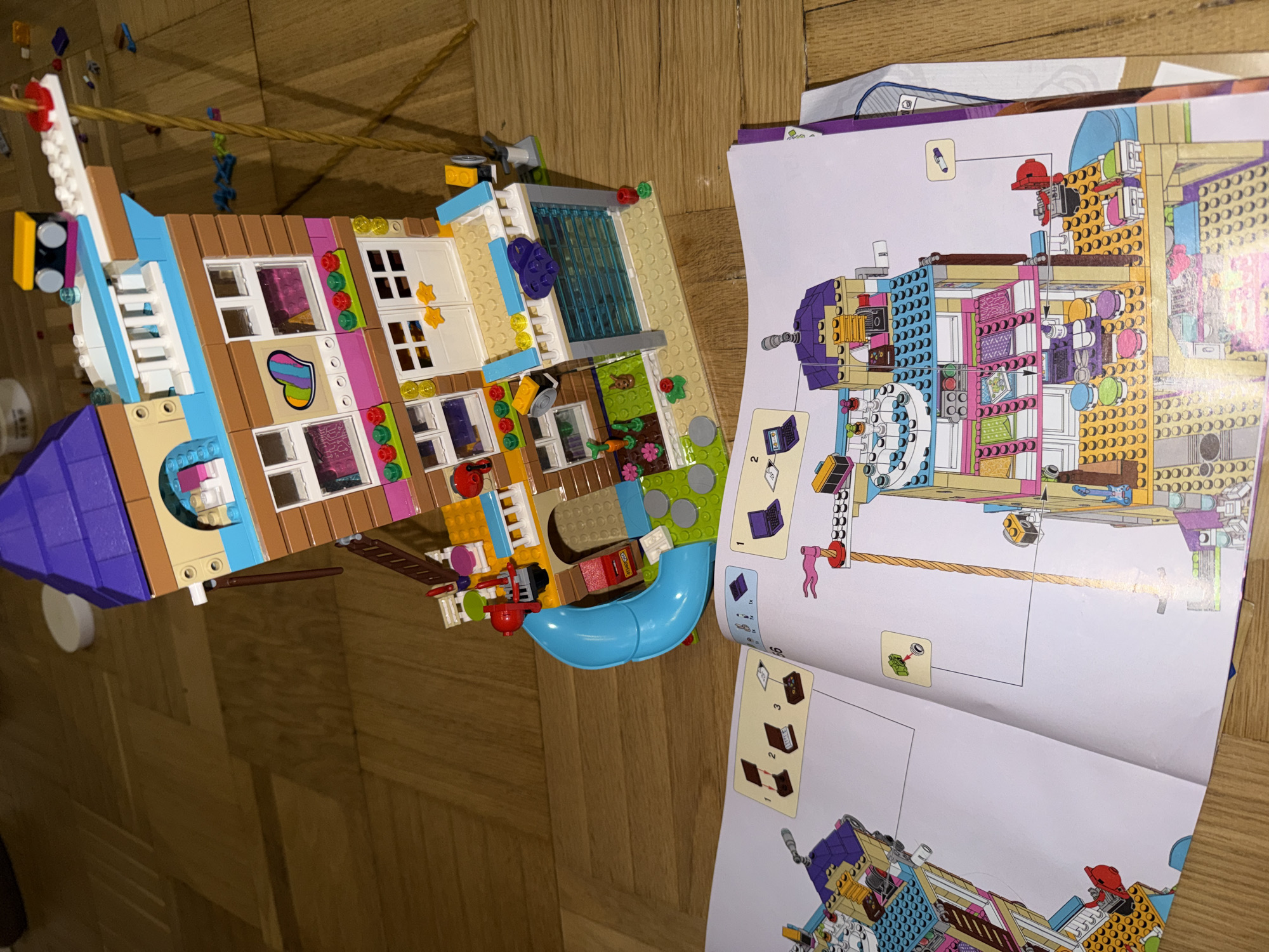 This is a top-down view of a partially assembled Lego set, specifically a Lego Friends building, resting on a wooden floor. The construction appears to be in progress, as evidenced by the Lego instructions manual open nearby. The set consists of multiple stories, with windows, doors, and other architectural details. A blue slide extends from one side of the structure. The open manual shows diagrams and step-by-step instructions for the assembly of the Lego set. The floor is made of wooden planks.