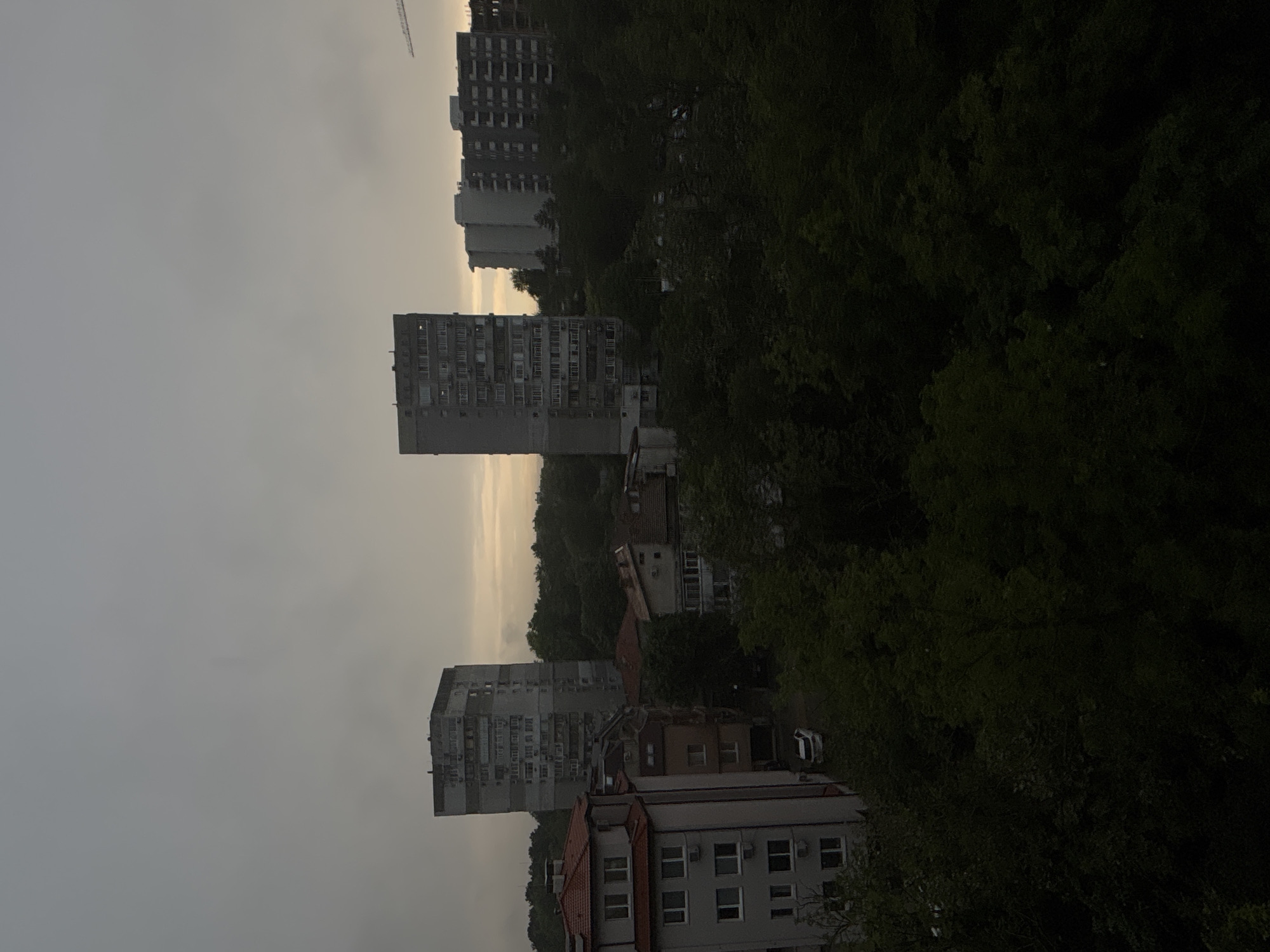 The image, rotated 90 degrees clockwise, captures an urban landscape characterized by multi-story residential buildings interspersed with dense green foliage and hilly terrain under an overcast sky. The sky is predominantly light grey, with a subtle band of lighter, pale yellow or orange light visible near the horizon, suggesting either a heavily overcast day, early morning, or late evening.
In the mid-ground, two large, grey, multi-story apartment blocks stand out, featuring numerous windows and balconies, some of which have air conditioning units attached. These buildings appear to be of an older architectural style, common in Eastern Europe. They are partially obscured and surrounded by a significant amount of dark green trees and bushes, which dominate the right side of the frame and cover the slopes between the structures.
In the foreground, towards the bottom right of the image, there is a building with a red-tiled roof, white-framed windows, and several air conditioning units, suggesting a more modern or recently renovated structure. A white car is partially visible near this building, tucked away among the trees. Further in the background, towards the upper left, another large building, possibly under construction or recently completed, is visible, along with a construction crane extending into the sky.
No specific activity or people are discernible. The scene presents a quiet, urban setting with a strong presence of nature. The muted lighting creates a calm, somewhat somber atmosphere. There is no visible text. The architecture and landscape are consistent with the provided location context of Varna, Bulgaria.