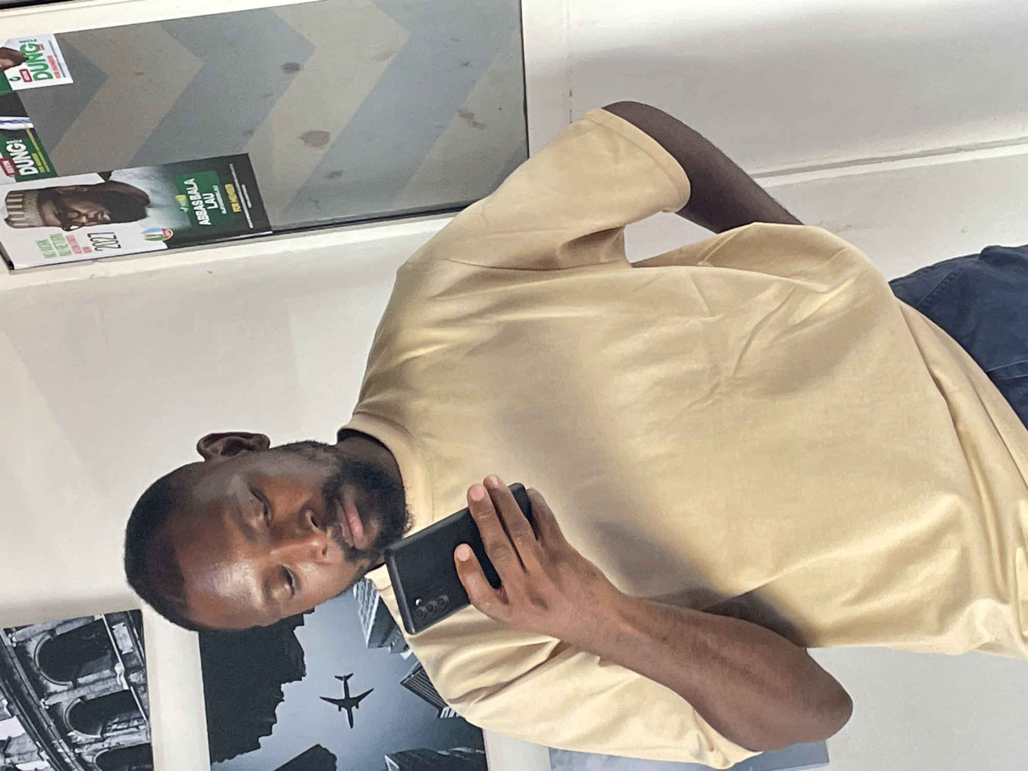 In Gura Top, Nigeria, a Black male stands indoors, holding a black smartphone in his right hand, his gaze directed downwards at the device. He is wearing a light tan t-shirt. In the background, a poster featuring a man in a cap and glasses, with text that includes "ABBAS BALA LAU" and "2027", is visible on a white wall. Another poster, partially obscured, also appears to be political in nature. To the left, artwork depicting architectural arches and a silhouette of an airplane is displayed on the wall. The environment appears to be a room with white walls and a window.