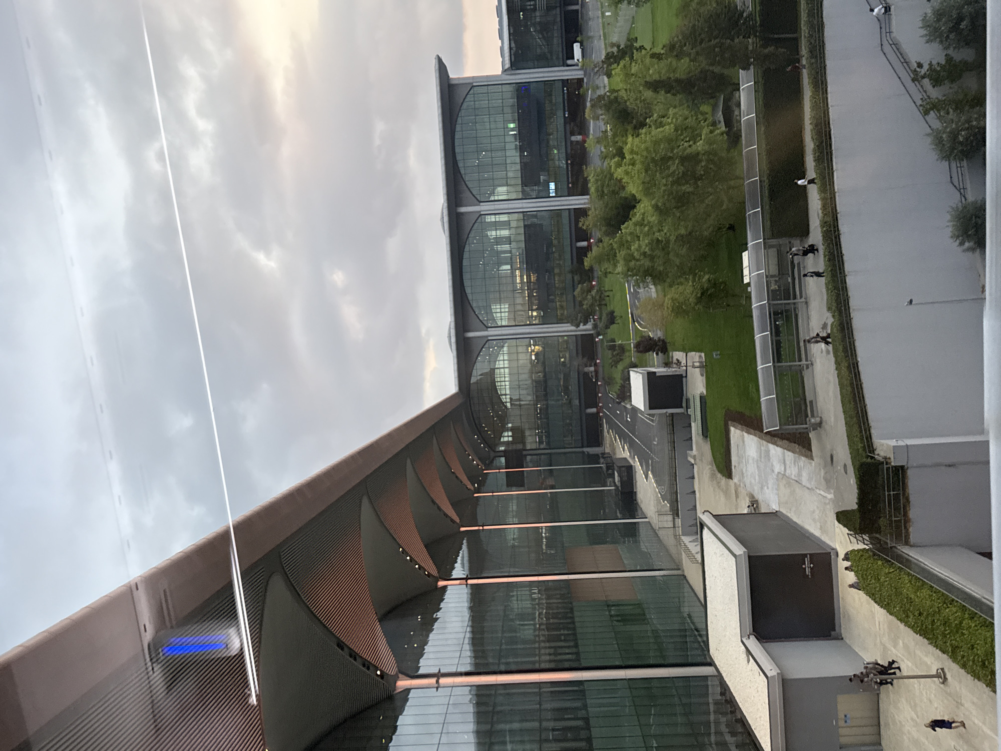The image shows a modern airport terminal with a large, curved, metallic roof and glass facade. The exterior is illuminated by artificial lights, casting a reddish-orange glow on the roof's underside. There is a blue light visible on the top left of the terminal. The sky above is overcast with scattered clouds, and a faint contrail is visible in the upper portion of the image.
In front of the terminal, there are paved walkways and grassy areas with trees. Several people are walking on the pathways. A white van is parked in the distance near the terminal's entrance. A road with traffic markings leads towards the building.
The lower portion of the image shows a multi-level structure with vegetation and a white retaining wall. Some individuals are visible on a walkway on this level, and a person is seen ascending a staircase.
The scene is consistent with an airport environment, likely in Arnavutköy, Turkey, given the presence of modern architecture often found in international airports. The time of day appears to be dusk or early evening, as indicated by the artificial lighting and the sky conditions.