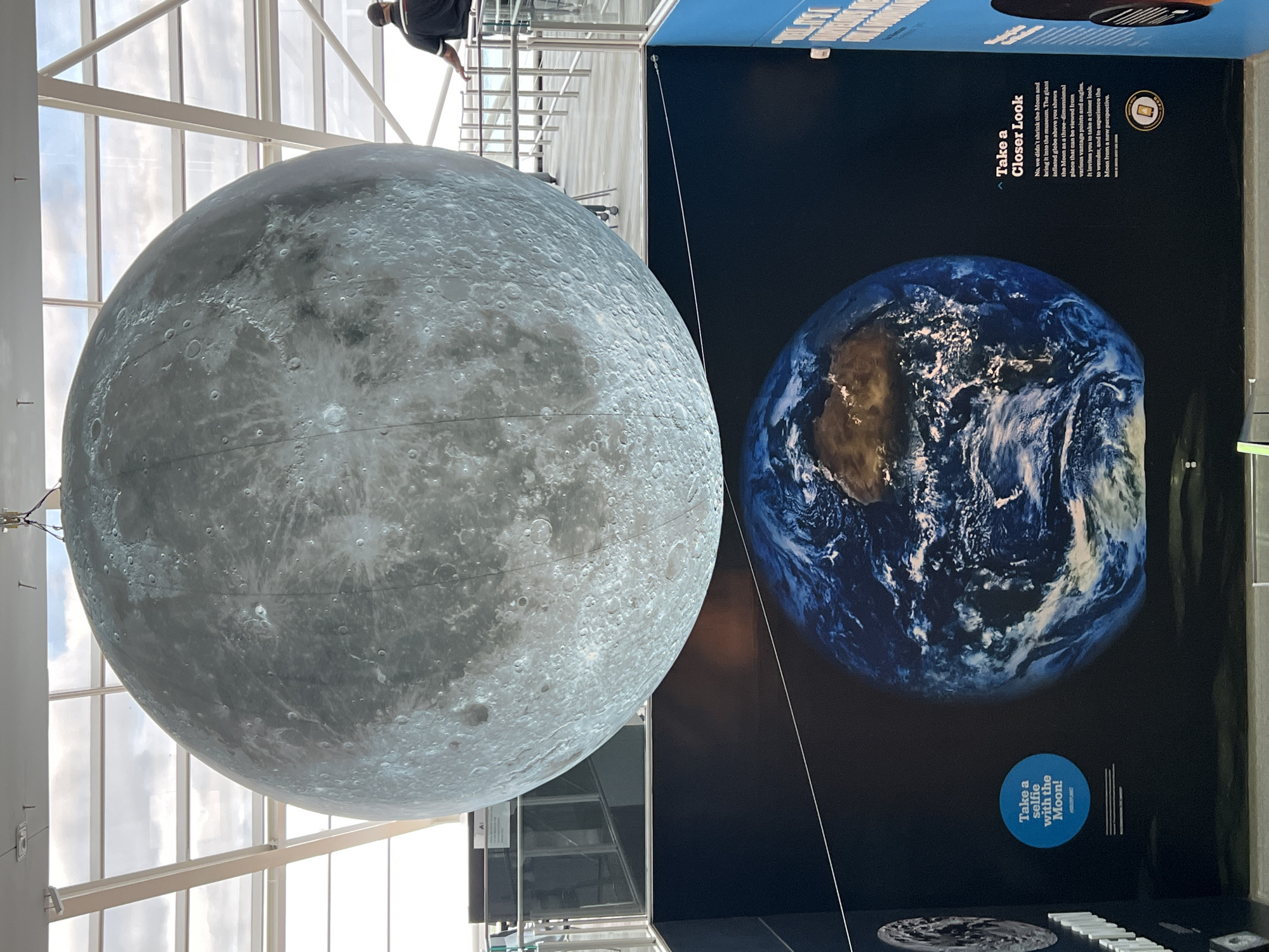 The image shows a spherical model of the Moon hanging inside a building with a large glass wall. The Moon model is gray and displays a textured surface covered with craters. Adjacent to the Moon model is a dark blue wall featuring a picture of Earth. Text on the wall reads "Take a Selfie with the Moon!" and "Take a Closer Look", followed by a paragraph explaining the exhibit. A person is partially visible at the top of the image, looking over a railing. The image was taken in Chicago, United States of America.