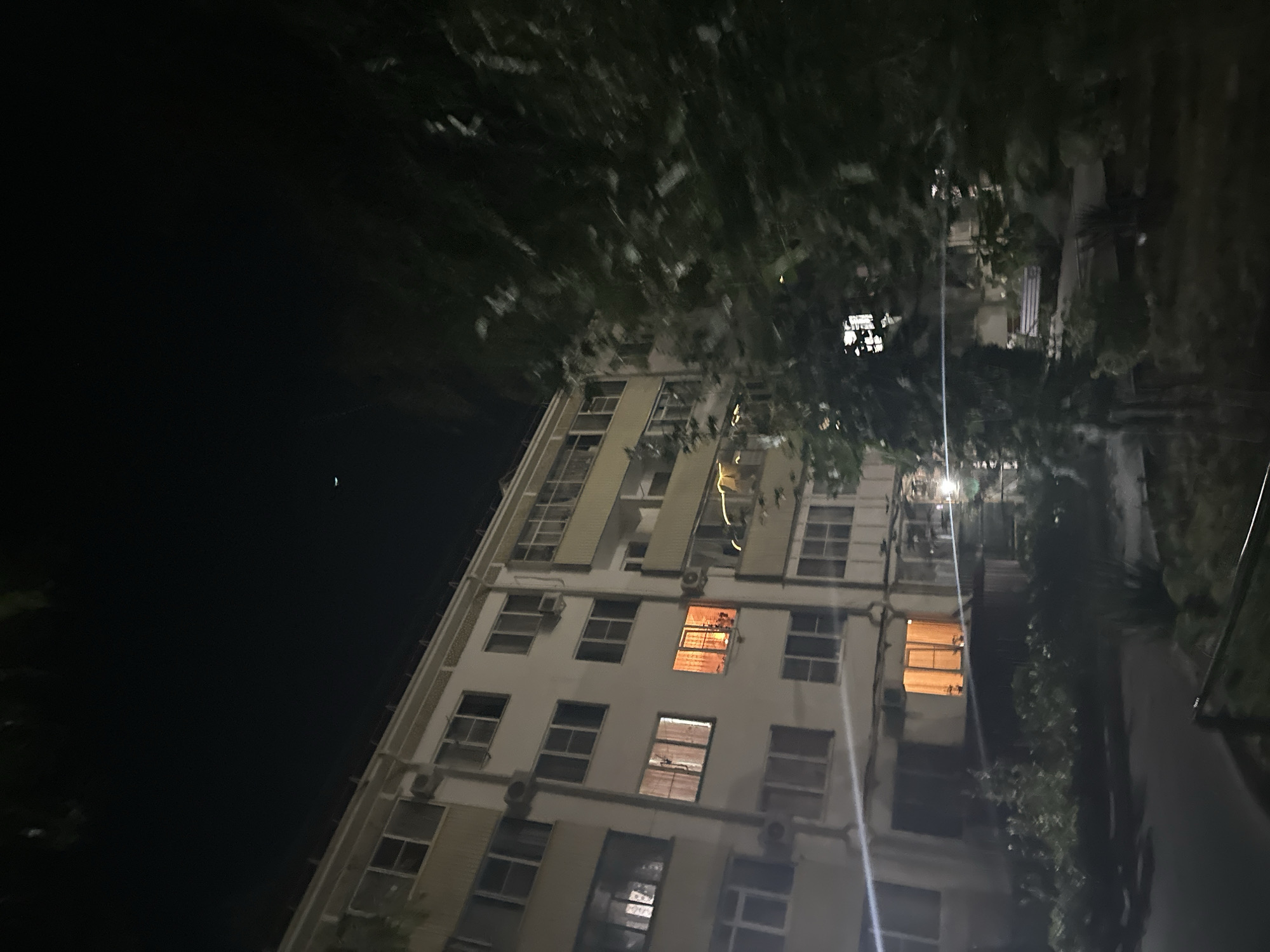 This is a nighttime exterior shot of a multi-story apartment building in the city of Nevinnomyssk, Russia. The building is constructed from light-colored panels and has multiple windows, some of which are illuminated from within, casting a warm orange glow. There are air conditioning units mounted on the exterior walls. In the foreground, there is a dark area with some foliage and a paved walkway. To the right, a large tree with dense foliage obscures part of the building. The sky above is dark, with a faint crescent moon visible. The scene suggests a residential area at night.