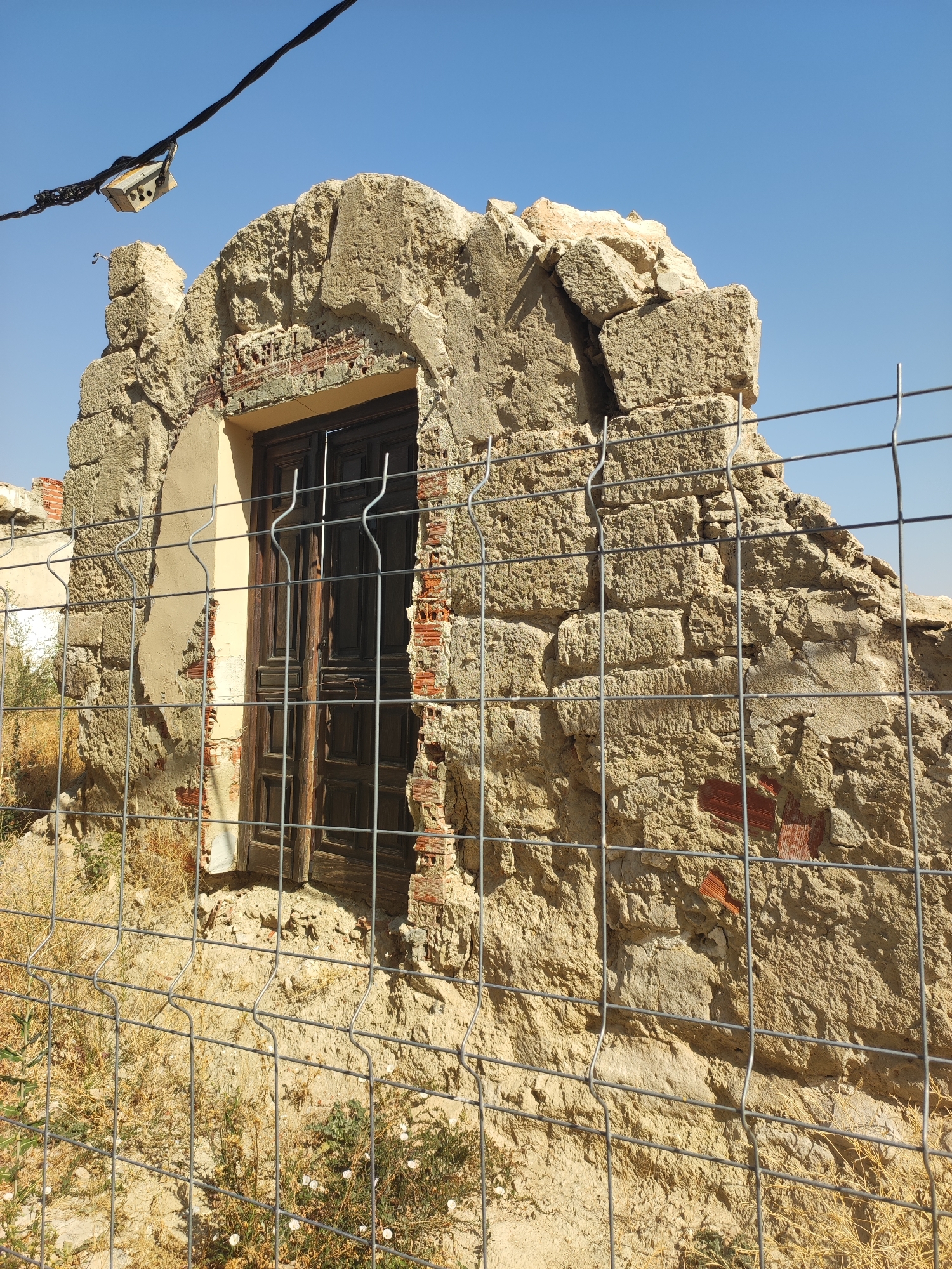 An outdoor, eye-level, medium shot captures a dilapidated stone building against a clear blue sky. A wire fence in the foreground obscures a weathered wooden door set into a crumbling stone facade. The building's construction is visible, with exposed red brickwork peeking through the rough-hewn stone. Dry, yellowed grass and scattered green foliage surround the structure. A black electrical wire with a small, light-colored box suspended from it stretches across the upper portion of the frame. The scene depicts an abandoned building located in Ampudia, Spain.