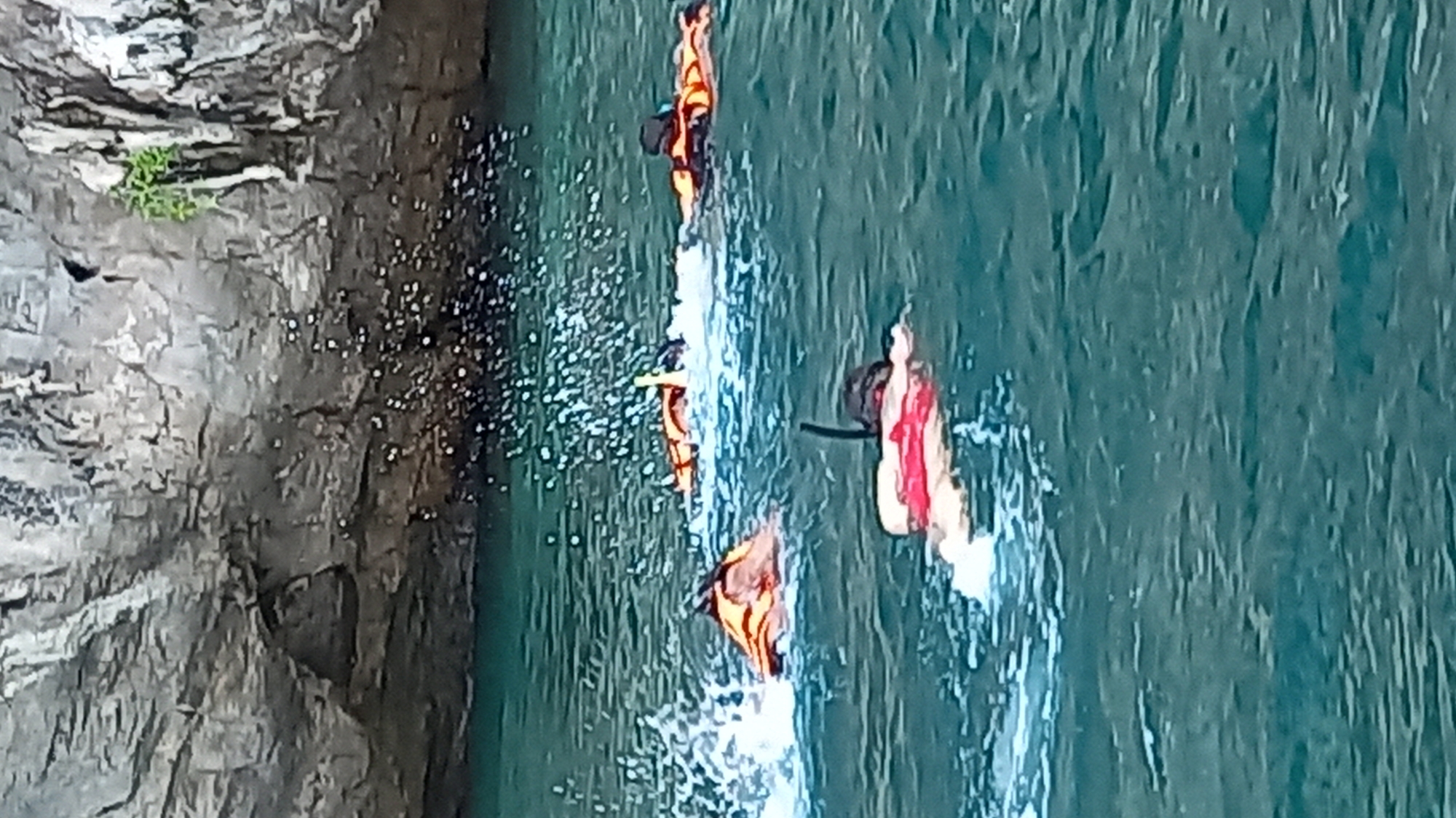 The image shows a cliff face on the left and water to the right. There are four people in the water wearing life jackets and swimming. The scene is in Ban Ao Nang, Thailand, based on the context.