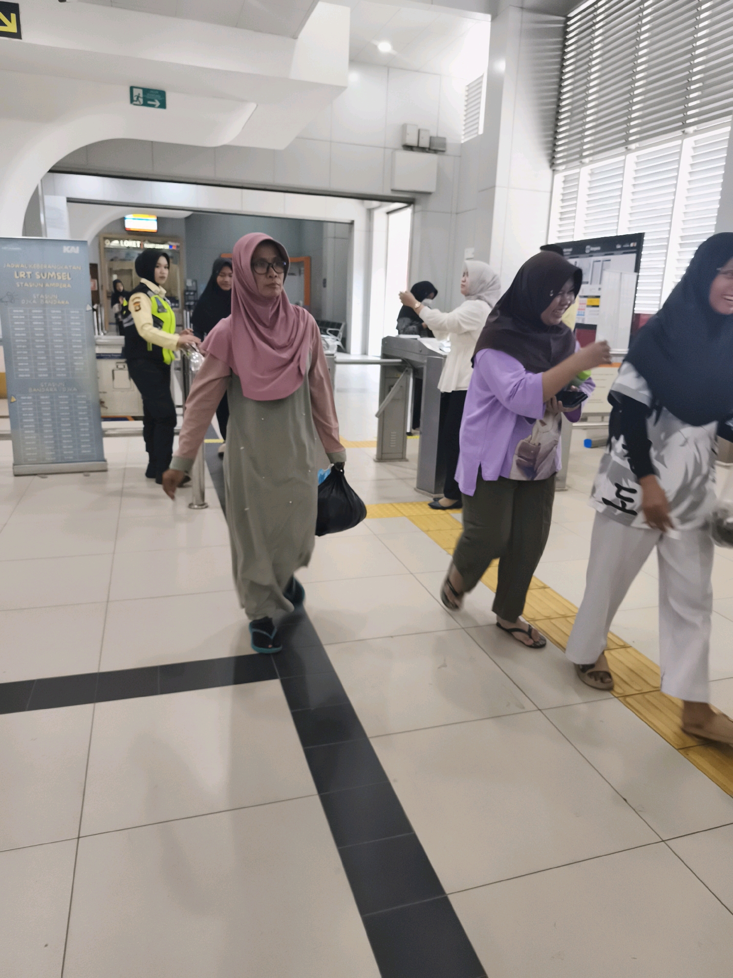The image shows an interior view of a public transportation station, likely a train or light rail station, in Kelurahan 16 Ilir, Kec. Ilir Timur I, Indonesia. The flooring is tiled with large square tiles, predominantly white with dark grey or black accent lines forming a grid pattern. In the foreground, three women are walking. The woman closest to the viewer is wearing a pink hijab, glasses, and a long, light grey dress with pearl-like embellishments. She is carrying a black bag in her right hand and has blue flip-flops on her feet. To her left, a woman in a brown hijab and a light purple top over dark olive pants is walking, holding a green bottle and a phone. To the far right, a woman in a black hijab and a white shirt with black patterns is walking, wearing white pants and sandals. In the background, a security officer in a yellow and black uniform is visible near a gate or turnstile. A large digital display board with schedules is mounted on a stand to the left, displaying "LRT SUMSEL" and "JADWAL KEBANGSAAN" (National Schedule). The environment appears modern and well-lit with fluorescent lighting and some natural light filtering in from a large window or skylight area with horizontal blinds in the upper right portion of the image. There are directional signs with green arrows on the wall in the background.