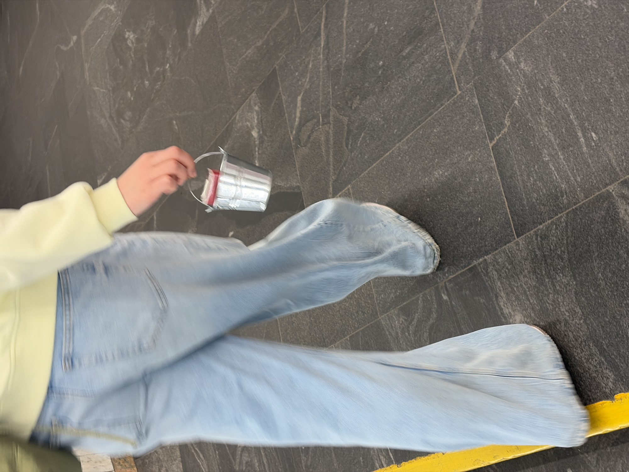 The image shows a person wearing light blue jeans and a pale yellow top, holding a small metal bucket with red and white items inside. The person is standing on a gray tiled floor with a yellow stripe running along the bottom of the frame. The photo appears to be taken from an overhead perspective. The location is not identifiable as Бараўлянскі сельскі Савет, Belarus based on the visual details provided.