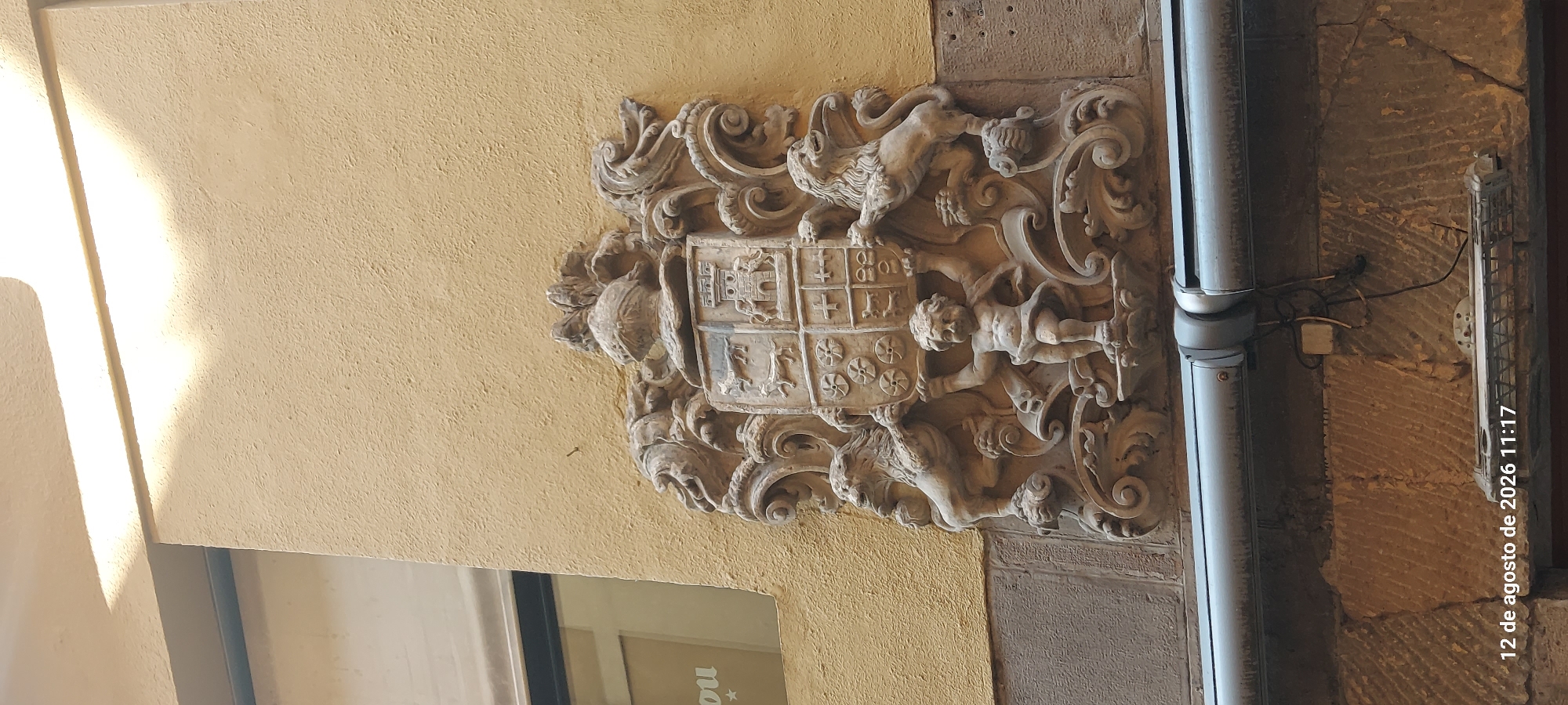 A close-up, low-angle outdoor shot captures a detailed, weathered stone coat of arms mounted on a textured, pale yellow building facade. The coat of arms is elaborate, featuring a central shield with multiple quadrants, adorned with architectural elements, animals, and a helmet at the top. Two rampant lions flank the shield, supporting it along with a cherubic figure positioned below. Ornate scrolls and floral motifs complete the decorative surround. A window with a partial reflection is visible to the left, and a metal gutter runs horizontally across the lower portion of the image, above rough-hewn stonework. The timestamp "12 de agosto de 2026 11:17" is superimposed on the bottom left corner. The scene is located on the facade of the Palacio de Navarra (Navarre Parliament Building) in Pamplona, Spain.