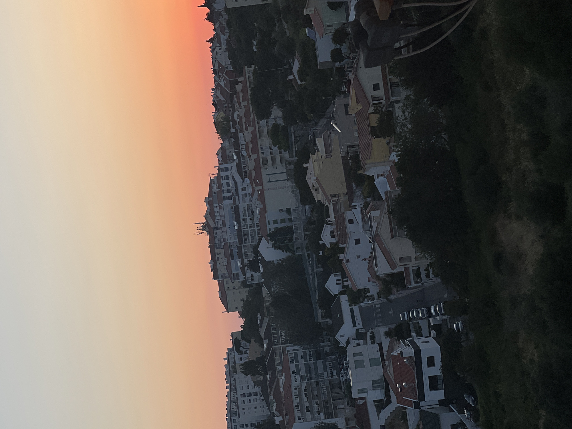 The image captures an elevated view of a residential area in Cascais, Portugal, during sunset. The sky displays a gradient of soft yellows, oranges, and pinks. The scene is dominated by a dense collection of buildings, predominantly houses with red-tiled roofs and multi-story apartment buildings. Trees and vegetation are interspersed throughout the urban landscape, with a darker, more densely wooded area in the foreground. A street is visible winding through the neighborhood, with several cars parked along its side. The overall impression is a tranquil, suburban setting bathed in the warm light of dusk.