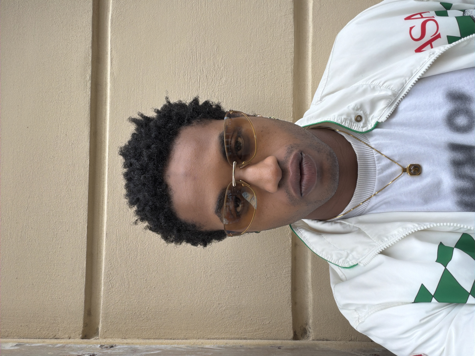 A close-up portrait features a young person, possibly male, with dark skin and dark, curly hair, looking directly towards the viewer. They are wearing gold-rimmed sunglasses with light amber-tinted lenses. A thin gold chain with a small, square, gold-framed pendant hangs around their neck, resting over a white ribbed t-shirt with faint, dark print. Over the t-shirt, they wear a white zipper jacket. The jacket has a green trim on the collar and features a green and white argyle pattern on the lower right side. Red capital letters spelling "ASA" are visible on the upper left side of the jacket. The background consists of a textured, light beige or tan wall, with a distinct vertical groove running through it. The lighting is bright and even, suggesting daytime outdoors. The subject maintains a neutral, direct gaze. The setting is contextually Paris, France.