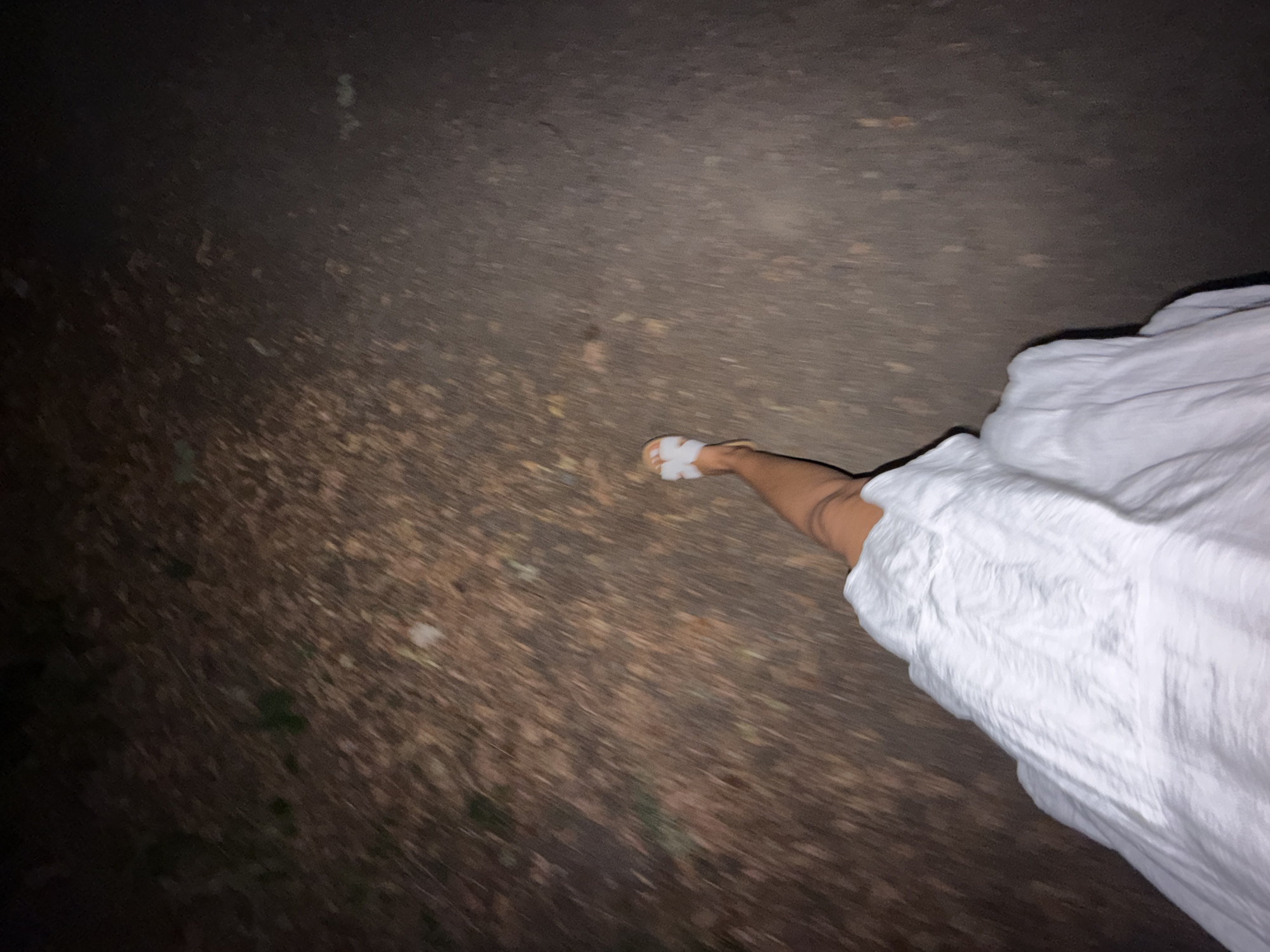 The image shows a first-person perspective of someone walking on a path covered with leaves. The person is wearing a white textured dress and white sandals. The path appears to be a dirt or gravel trail, with some patches of darker, possibly wet, ground. The lighting suggests it is nighttime or very early morning, with limited visibility. The scene is outdoors, likely in a park or wooded area, given the presence of leaves. The location is identified as Bruchköbel, Germany, but no specific landmarks are visible to confirm this. The motion blur indicates the person is in motion.