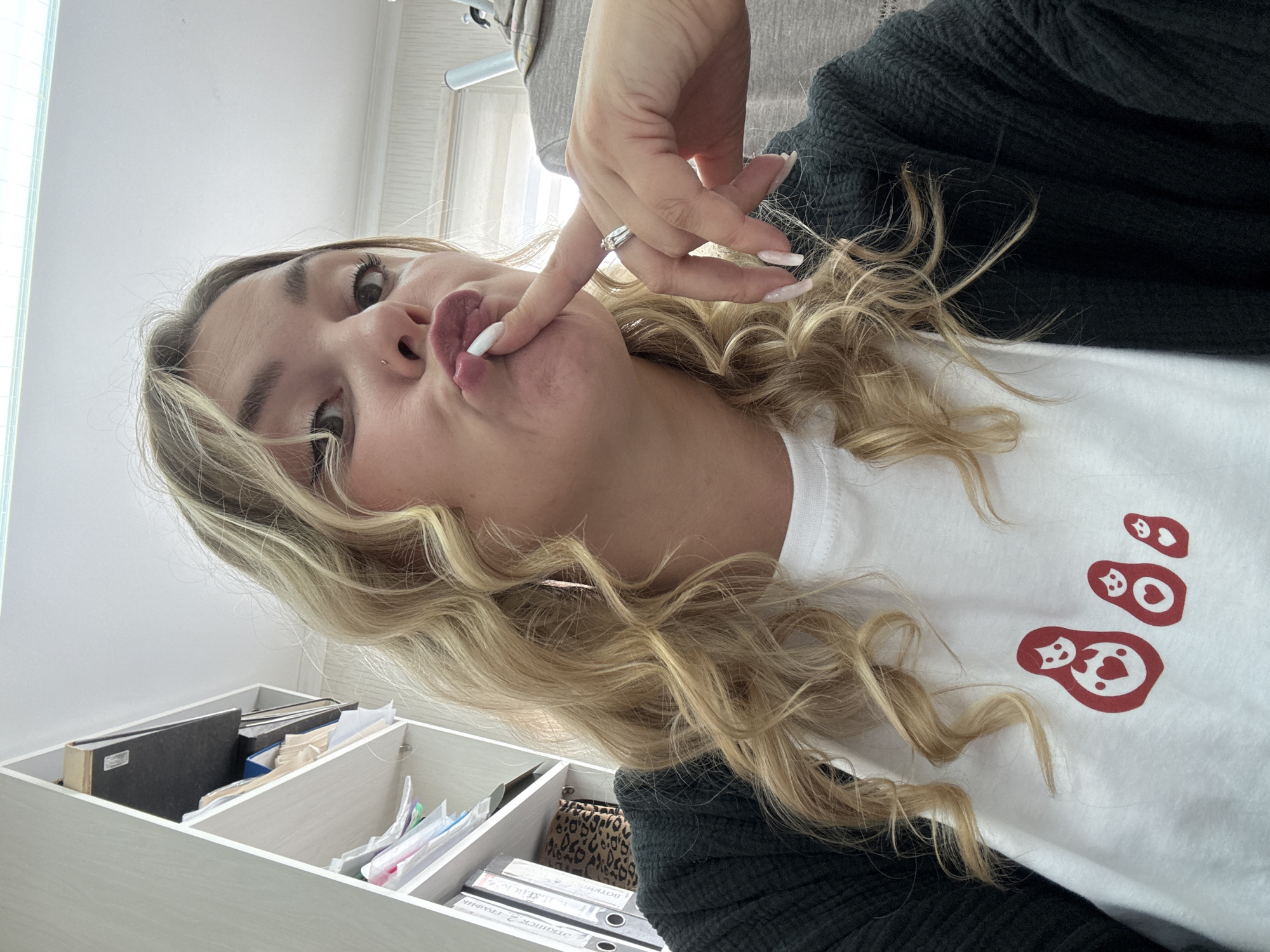 In a medium shot, a young woman with blonde, wavy hair is captured from the chest up, looking towards the camera with her lips puckered. She is wearing a white t-shirt with a red graphic of nesting dolls and a dark green cardigan. Her right hand is raised, with a finger touching her lips. She is wearing a ring on her right ring finger and has long, manicured fingernails. In the background, a white shelf unit is visible, filled with binders and papers. The lighting suggests an indoor environment, possibly an office or home. The image is taken at an upward angle, and the overall impression is a candid selfie. The specific location in Noviy, Russia, cannot be determined from the visual information provided in the image.