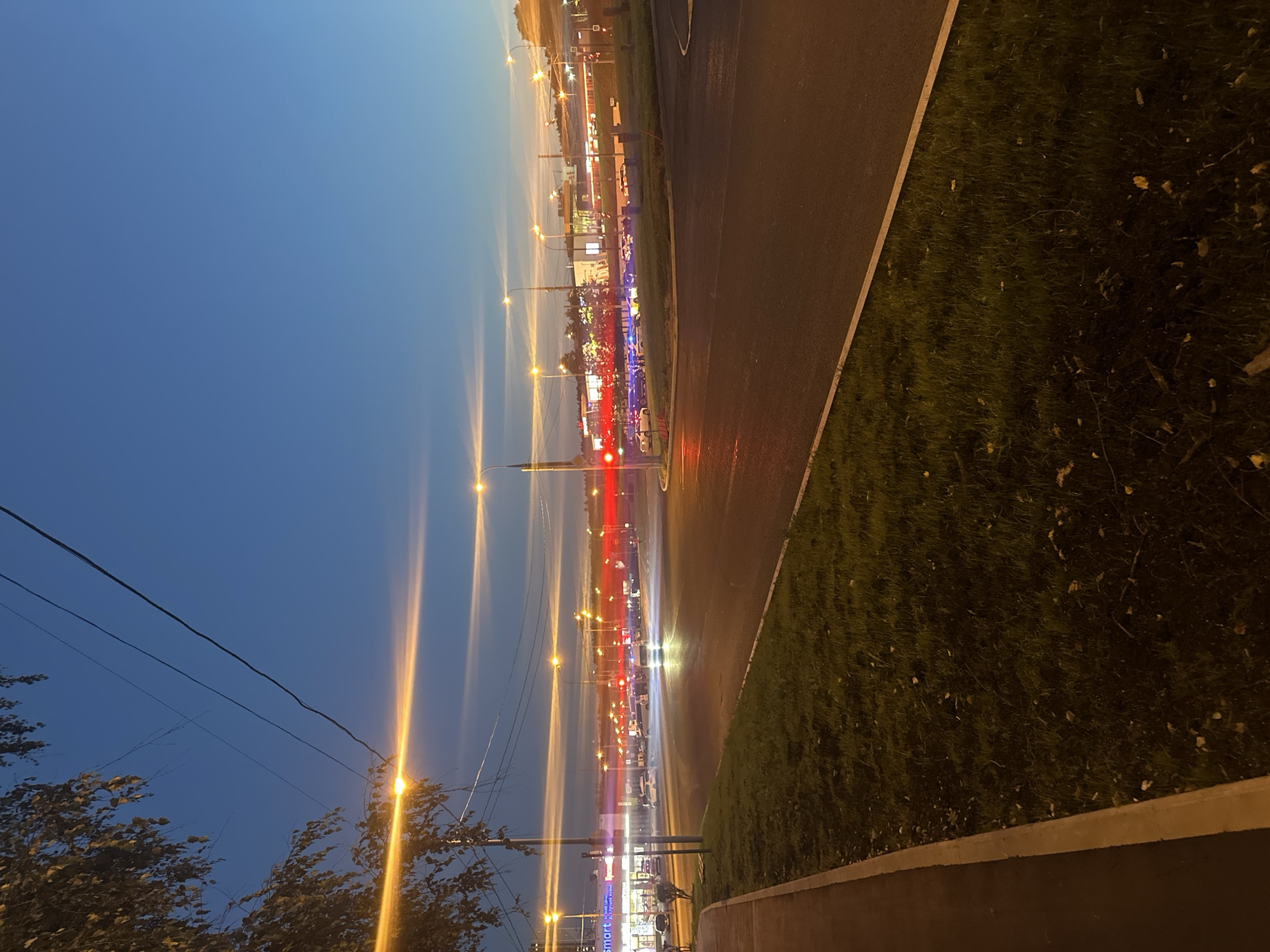 The image depicts a traffic intersection at dusk, possibly in Moscow, Russia, as indicated by the presence of Cyrillic script on a store sign ("smart"). The sky is a deep blue with wispy clouds illuminated by the setting sun. Streetlights are casting long, bright orange beams, and the headlights and taillights of passing vehicles have created light trails due to a long exposure.
In the foreground, a sidewalk borders a grassy area. The road itself is wet, suggesting recent rain. In the midground, cars are navigating the intersection. On the left, a building with a prominent "smart" sign suggests a commercial area, possibly a supermarket or retail complex. Further down the road, more buildings and streetlights are visible. The overall atmosphere is urban and slightly blurred due to the motion of traffic.