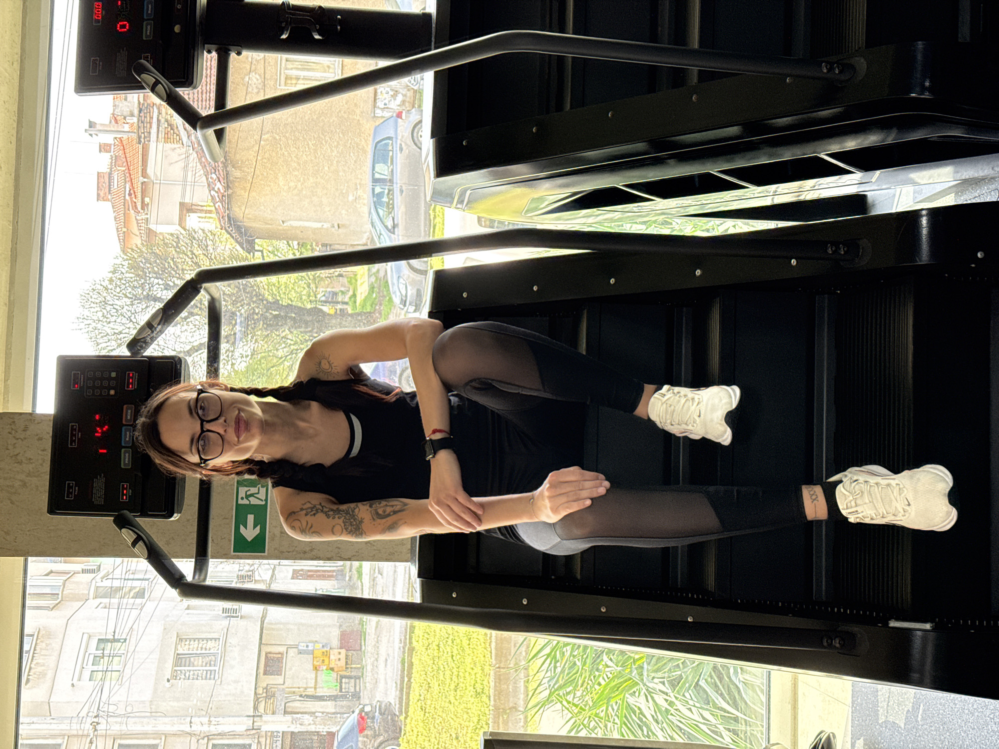 A woman with glasses and pigtails is sitting on a stair climbing machine in what appears to be a gym. She is wearing a black top, leggings with mesh panels, and white sneakers. Her arms are resting on her legs. There are tattoos visible on her arm. Outside the window, buildings are visible, suggesting an urban environment. A green emergency exit sign is present. Based on the architecture and the outdoor scene, it is likely that the location is in the city of Pleven, Bulgaria.