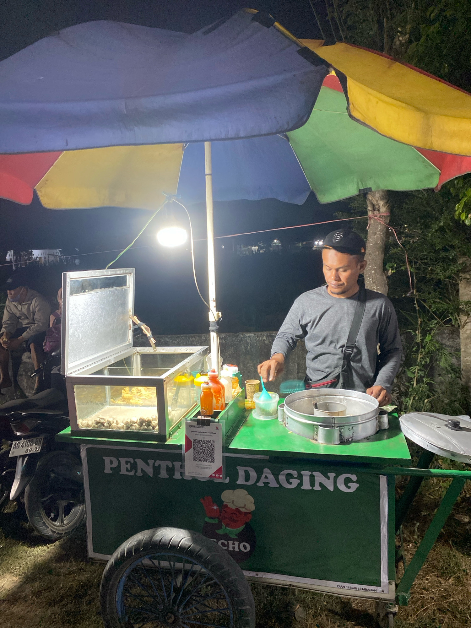 A man is preparing food at a food cart in Branggahan, Indonesia. The cart is green with a colorful umbrella canopy. The man is wearing a grey long-sleeved shirt and a black baseball cap. He is standing behind the cart, holding a blue utensil and interacting with a metal container. In front of him, there is a glass display case filled with food items, and several bottles of condiments. A motorcycle is parked to the left of the cart. The scene appears to be outdoors at night, with a tree and foliage visible in the background.