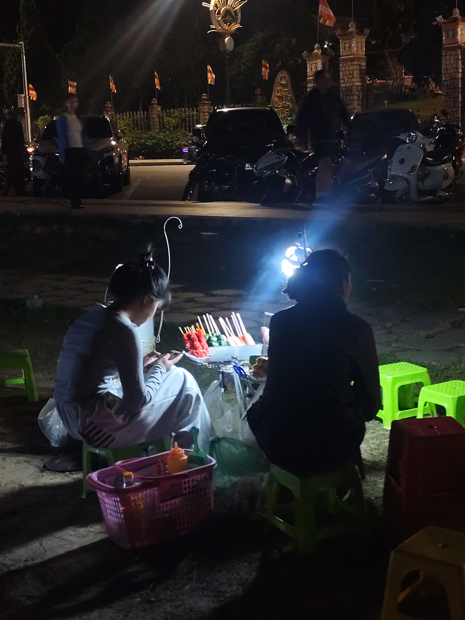 The image depicts a nighttime scene on a street in Da Lat, Vietnam, likely near a tourist area or market. In the foreground, two individuals, appearing to be women, are seated on low plastic stools. One woman, facing away from the camera, is seated on a green stool. The other woman, also facing away from the camera, is seated on a green stool and appears to be looking at a smartphone. In front of the women, a basket contains bottles of condiments. Various food items on skewers are displayed on a small table. The background features several parked motorcycles and scooters, some covered with tarps, suggesting a vehicle parking area or a street vendor setup. The lighting is dim, with artificial lights from street lamps and possibly food stalls illuminating the scene. The presence of flags and architectural elements suggests a cultural or religious setting. The overall atmosphere is that of a busy night market or a roadside eatery.