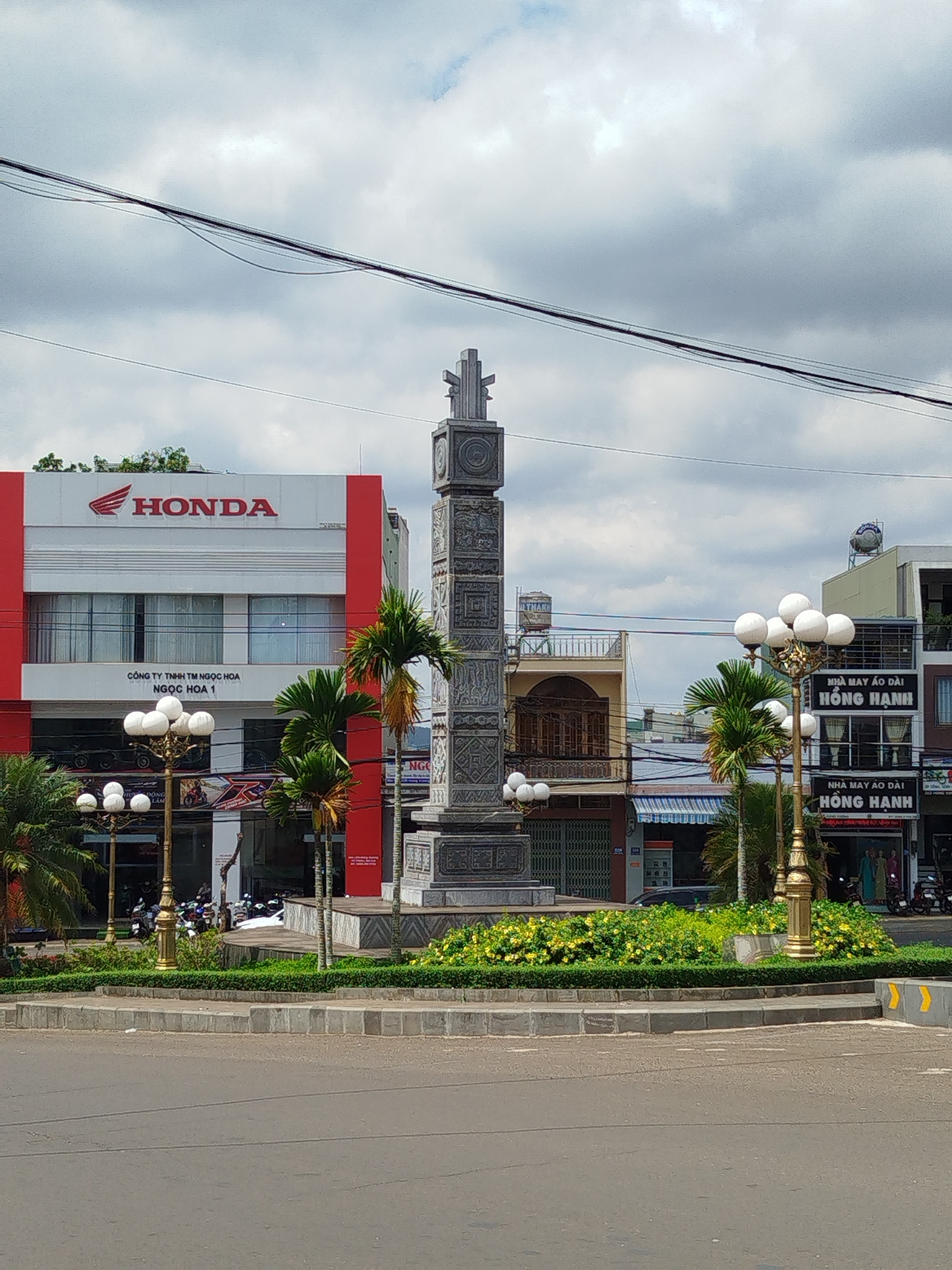 The image depicts a street scene in Pleiku Ward, Vietnam, featuring a roundabout with a tall, ornately decorated stone monument in the center. Lush greenery and flowering plants surround the base of the monument. Streetlights with multiple spherical bulbs flank the monument on both sides.
In the background, buildings line the street. On the left is a red and white Honda dealership, with the Honda logo prominently displayed. To the right are other commercial buildings, including one with the sign "NHÀ MAY ÁO DÀI HỒNG HẠNH," which translates to "Hồng Hạnh Áo Dài Tailor Shop." The sky is overcast with heavy clouds. Power lines are visible overhead. The street is paved, and there are curbs along the edge of the roundabout. There are multiple scooters parked in front of the Honda dealership and tailor shop.