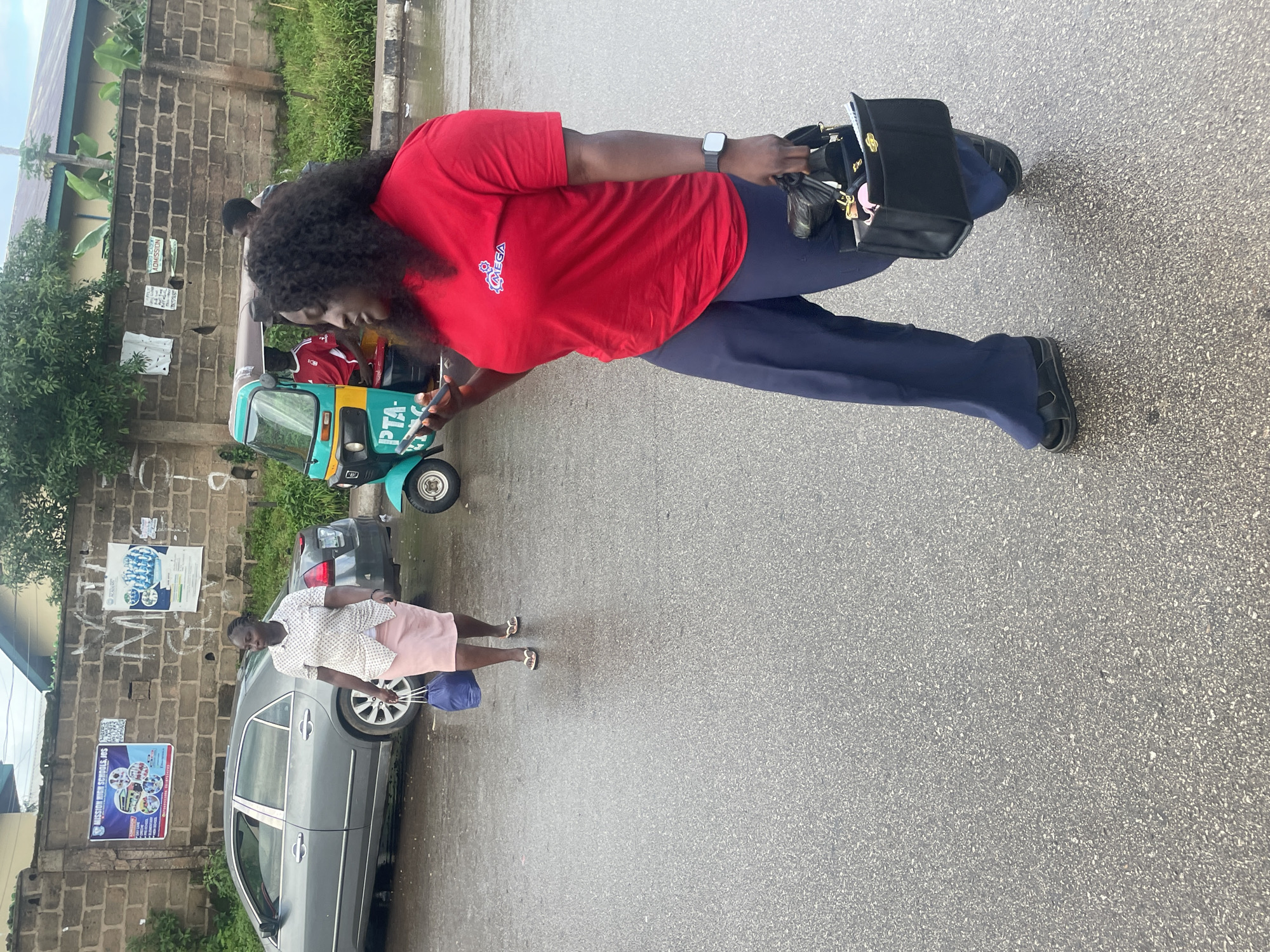 The image shows a street scene in Barakin Lerawi, Nigeria. In the foreground, a Black woman wearing a red t-shirt and dark blue pants walks towards the right, holding a black handbag and a mobile phone. She is wearing black sandals and a smartwatch. Behind her, a light-skinned woman wearing a white polka-dot blouse and a pink skirt walks towards the left, carrying a blue tote bag. She is wearing flip-flops. Between the two women, a turquoise auto-rickshaw is parked. The ground is wet, suggesting recent rain. In the background, a grey sedan is parked next to a brick wall with posters on it. Trees and vegetation are visible behind the wall, and the sky is overcast.