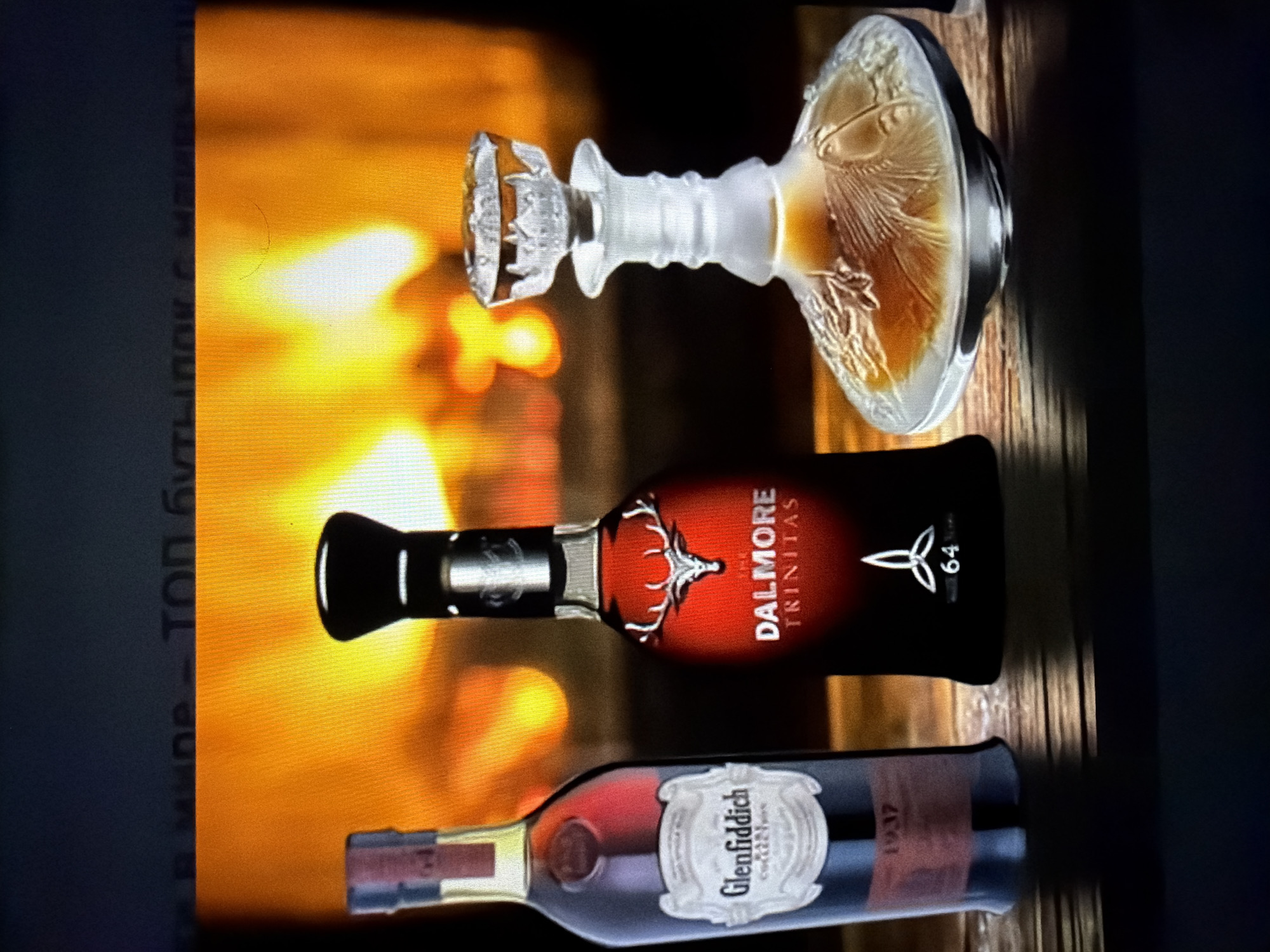 In the city of Falenty Nowe, Poland, three bottles of alcohol and a crystal decanter sit on a wooden surface. Behind them, a fireplace glows with warm, orange light. The bottle on the left is labeled "Glenfiddich." The bottle in the middle is dark red and black with "The Dalmore Trinitas" written on it, along with a stag's head emblem. The crystal decanter on the right is filled with amber liquid. The background is a blurry depiction of a roaring fire.
