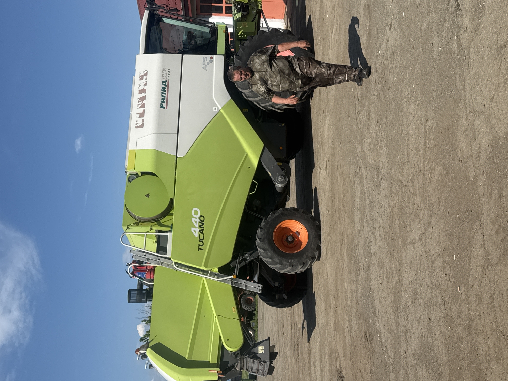 The image depicts a man standing in front of a CLAAS TUCANO 440 combine harvester, presumably in Sarnevo, Bulgaria. The combine harvester is a light green and white color. The man is wearing camouflage clothing. The ground is a concrete or paved surface, and the sky is visible with a blue sky.
