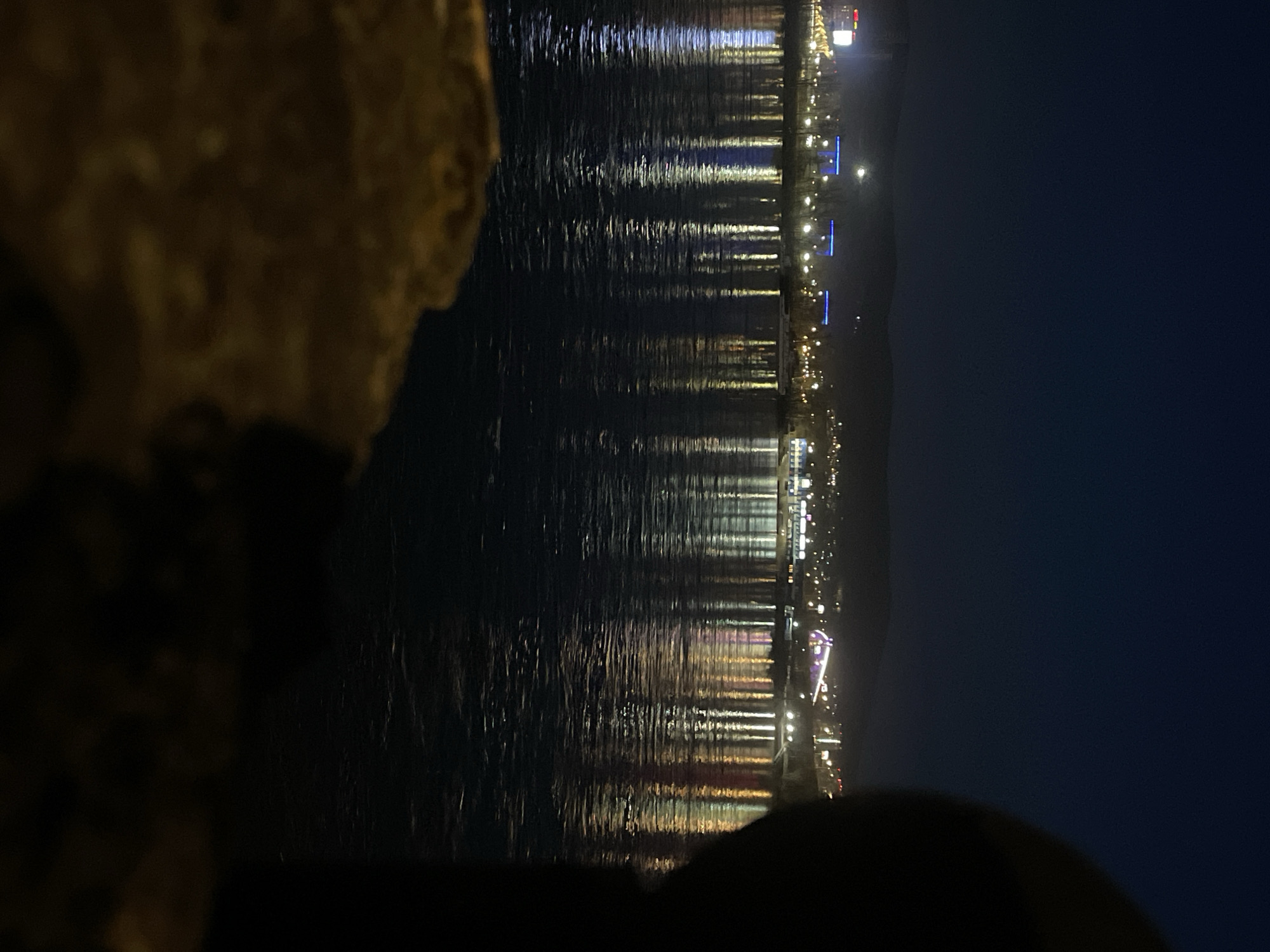 The image displays a nocturnal scene overlooking a body of water, likely in Krasnoyarsk, Russia, with a cityscape illuminated on the opposite bank. The perspective suggests the photograph was taken from a bank or an elevated position overlooking the water.
In the immediate foreground, a large, blurry, dark, and irregularly shaped object, possibly a rock or part of a structure, occupies the left side, partially framing the view.
The central part of the image is dominated by the dark surface of the water, which reflects the numerous lights from the distant shore. These reflections appear as elongated, shimmering vertical streaks, indicating slight movement on the water's surface. The reflected lights exhibit a variety of colors, predominantly bright white and warm yellow from streetlights and building illuminations, along with distinct blue lights and occasional hints of red and purple.
On the far bank, a continuous line of urban lights stretches across the horizon, marking the presence of buildings and structures along the waterfront. These lights create a vibrant glow against the dark night. Beyond the illuminated cityscape, the background is a dark, indistinct mass, likely representing distant hills or the unlit portions of the landscape merging with the night sky.
The time of day is clearly night. No specific activity or event is taking place, and no visible text can be discerned in the image. The overall atmosphere is peaceful and urban at night.