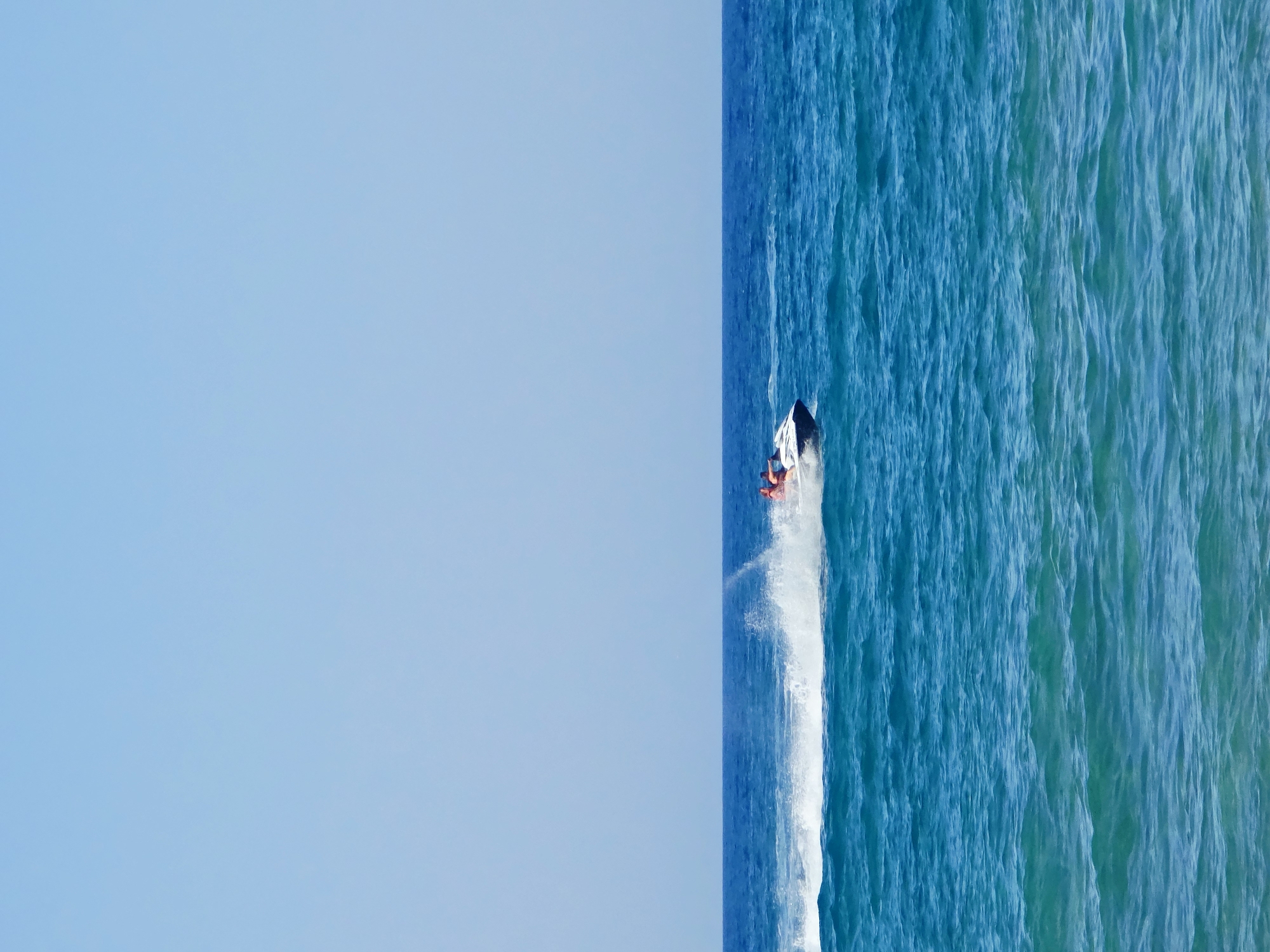 A jet ski is seen in motion on the sea, creating a white wake. The jet ski is carrying two individuals, both wearing orange life vests. The water is a deep blue, with some lighter turquoise waves visible in the foreground. The sky above is a clear, pale blue. The scene appears to be located in the open sea off the coast of Malia Municipal Unit, Greece, with no land visible in the frame.
