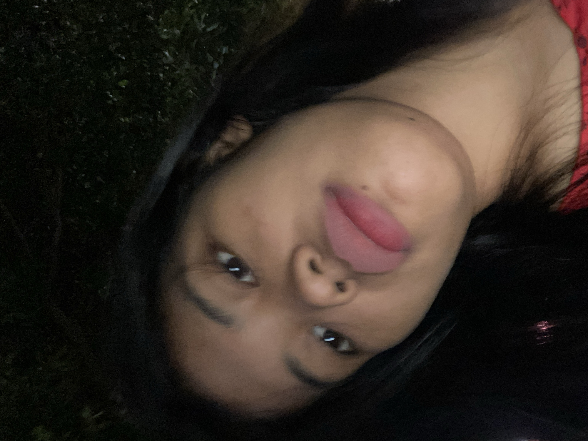 A close-up, selfie-style photograph shows a young woman with dark hair and brown eyes looking directly at the camera. Her mouth is slightly open, and her tongue is visible, extending towards her upper lip. The background is dark and appears to be outdoors, with some blurred green foliage visible. The image is taken at night, and the lighting is dim, creating a slightly grainy texture. The woman is wearing a red garment, part of which is visible at the bottom of the frame. The image is a candid shot, likely taken by the subject herself. The location of the image is not specified, and therefore, it is not possible to determine if it is in Madiun, Indonesia.
