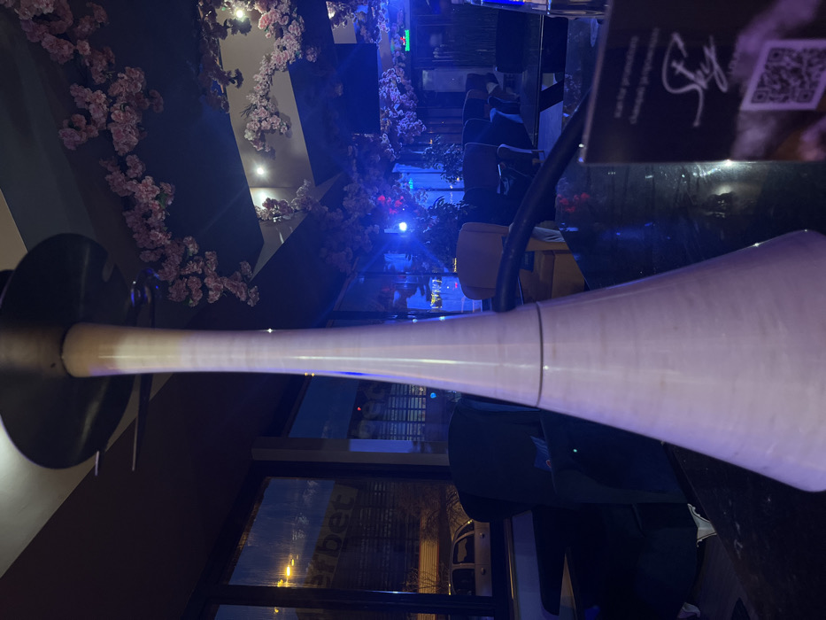The scene depicts the interior of a lounge or shisha bar at night, confirmed to be in Sofia, Bulgaria, by a visible "efbet" sign through the exterior windows. A tall, conical, light-colored modern hookah stands centrally in the foreground on a dark, reflective tabletop. To the right of the hookah, a partially visible menu card displays the text "Sky" and a QR code. The lounge interior features a seating arrangement with dark-toned and some lighter beige/yellow armchairs. The ceiling is decorated with extensive garlands of artificial pink cherry blossoms, illuminated by ambient blue lighting. Large windows in the background offer a view of an urban street at night, showing streetlights, reflections, and a white parked vehicle. No individuals or explicit actions are visible within the frame.