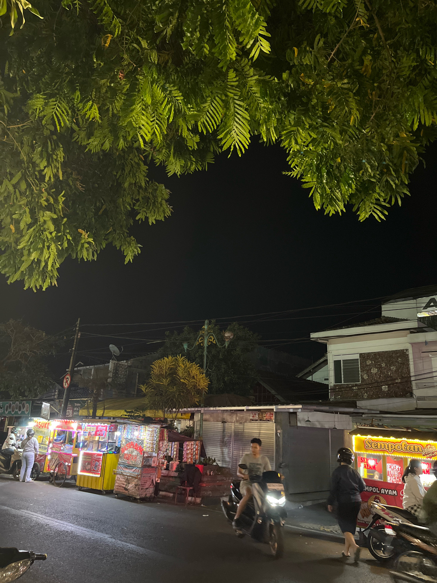In Salatiga, Indonesia, at night, the scene unfolds on a street lined with illuminated food stalls. Overhead, lush green leaves of a tree cast dappled shadows. The stalls are a vibrant spectacle of light and color, displaying an array of goods. One stall, brightly lit in yellow, advertises "Sempolan Ayam," with signs mentioning "Sedeap Alam" and "Everything Rich." Next to it, a smaller stall, also illuminated, features a variety of products displayed on shelves. Another stall, predominantly yellow with red accents, shows a red bicycle wheel and further merchandise.
On the asphalt road, several people are engaged in nighttime activities. A man rides a scooter towards the viewer, his headlight illuminated. Another scooter is parked nearby. Two individuals stand near the yellow and red stall, one appearing to be a vendor. Further down the street, a person walks away from the "Sempolan Ayam" stall, dressed in dark clothing and a helmet. Another scooter is parked in front of this stall, with a person on it wearing a helmet. A woman in a light-colored jacket stands near the "Sempolan Ayam" stall. A street lamp with an ornate design stands tall on the right side of the image. In the background, buildings with tiled roofs and windows are visible, along with utility poles and wires, indicating an urban environment. A satellite dish is mounted on a pole in the distance.
