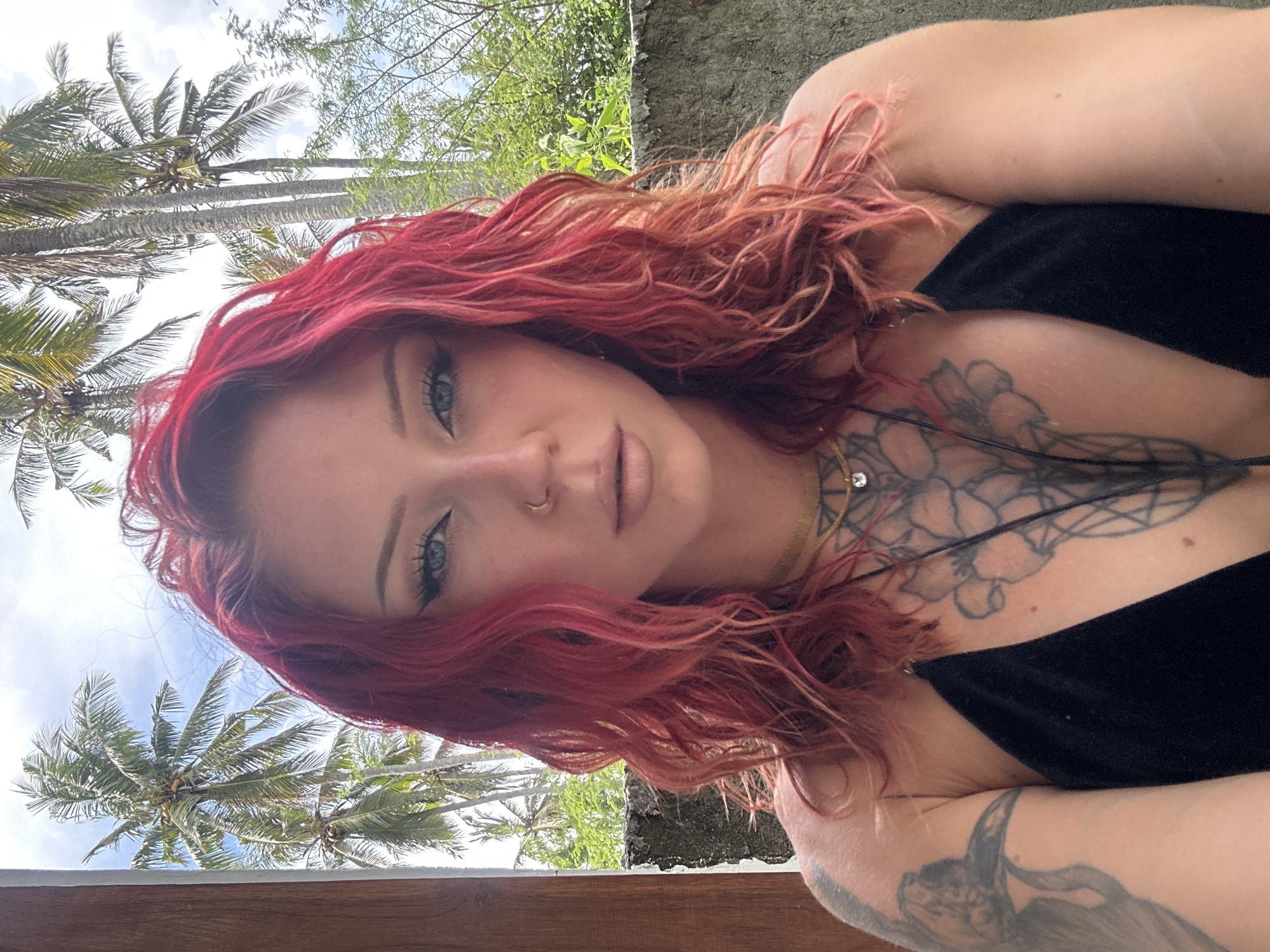 A woman with red, wavy hair and tattoos on her chest and arm is lying down, with the backdrop of palm trees in Nusa Penida, Indonesia. She is wearing a black top. The sky is visible through the trees.