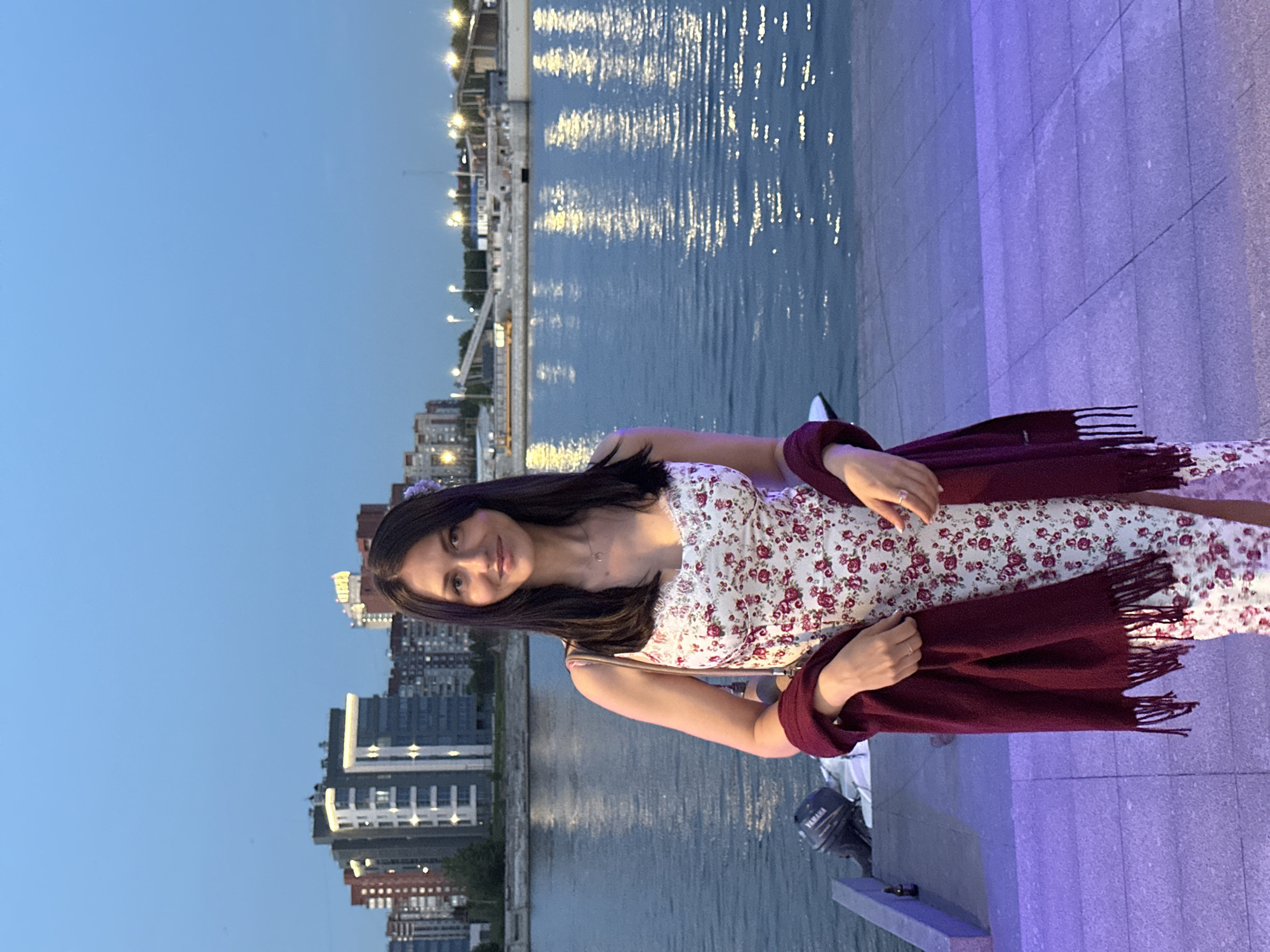 A young woman stands on a paved walkway next to a body of water in Saint Petersburg, Russia. She is wearing a white floral dress and a burgundy scarf draped over her shoulders. In the background, the lights of the city buildings are visible across the water. The sky is a deep blue, suggesting it is dusk. A small boat with a Yamaha motor is docked to her left.