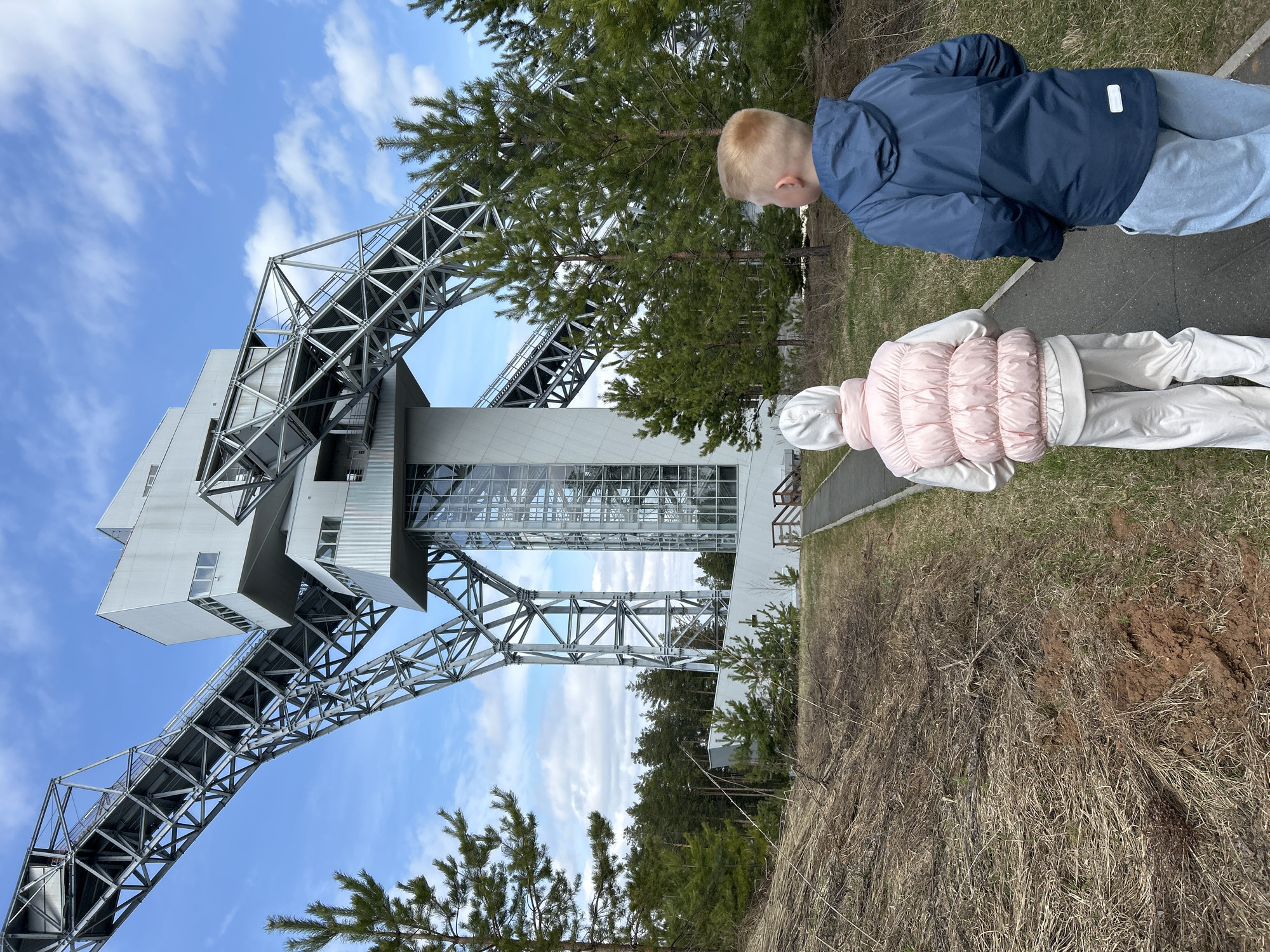 A ground-level, wide-angle shot shows two children on a sidewalk in front of the Ski Jumper Training Center, an athletic facility in Прикамский, Russia. The structure's design features an elevator tower and connecting bridge. To the right of the frame, a young boy with short blonde hair wears a blue jacket. Next to him, a girl wearing a white tracksuit and a pink puffer vest faces away from the camera. Sparse, dry vegetation and a few evergreen trees fill the midground and background, while a partly cloudy sky is visible above the structure.