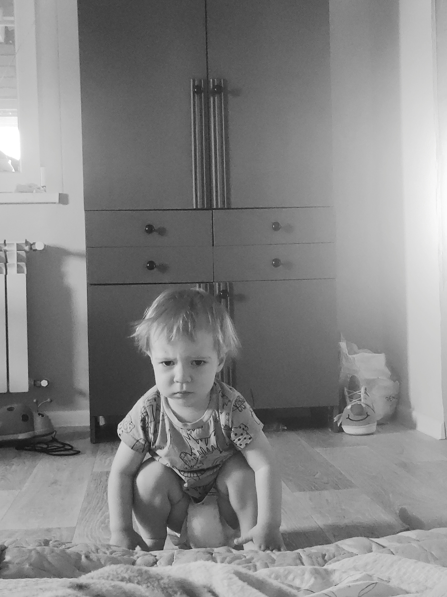 A black and white medium shot depicts a young child, appearing to be a toddler, squatting on a blanket or mat on a wooden floor. The child has fair hair and a stern expression. Behind the child is a tall wardrobe or cabinet with two doors on top and two rows of drawers below, with simple round knobs. To the left of the cabinet is a white radiator near a window. In the background, to the right of the cabinet, there's a pair of shoes. The location is a room in a house or apartment, in an unknown city.