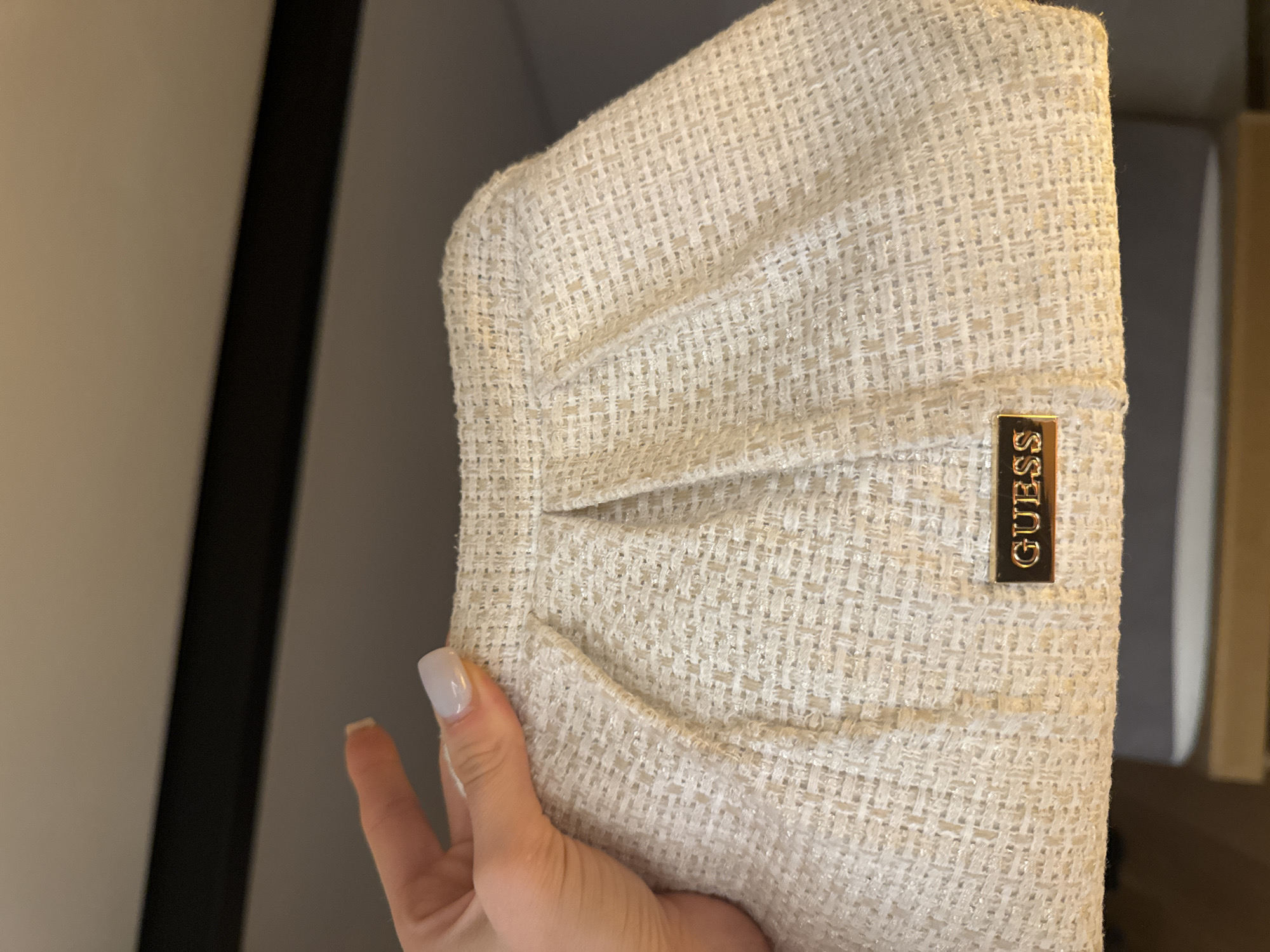 A close-up, top-down shot showcases a cream-colored textured clutch bag held by a woman's hand with light-colored, manicured nails. The bag features subtle pleats running vertically down its front and a gold-toned rectangular plaque with the embossed word "GUESS" near the bottom edge. The background is out of focus, suggesting an indoor setting. The scene is devoid of any recognizable landmarks of Murcia, Spain.