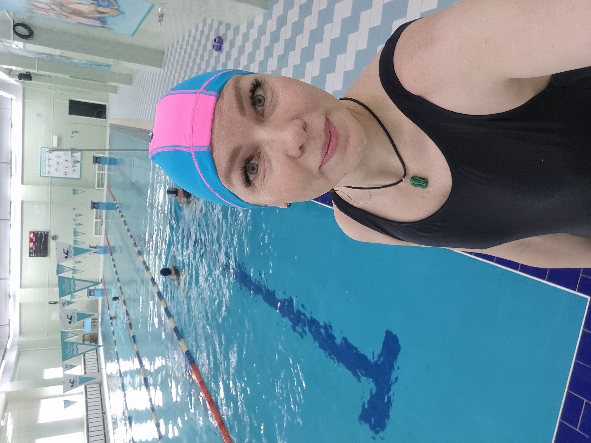 A woman wearing a black bathing suit and a pink and blue swimming cap stands in a swimming pool at what appears to be a municipal pool. Several swimmers are in the background lanes of the pool, which is marked with lane ropes and T-markers at the ends. A large scoreboard is visible. The pool area is tiled in blue and white. She is wearing a necklace with a green pendant. The location is Markovskii, Russia.