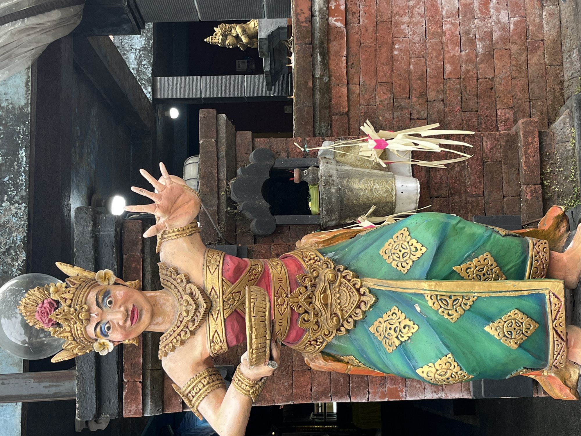 A full-body, eye-level, medium shot captures a colorful, life-sized statue of a Balinese dancer standing in front of a red brick wall in Kuta, Indonesia. The statue is elaborately adorned with gold accents and a vibrant green and red sarong. The dancer has striking blue eye makeup and a golden headdress adorned with a pink flower. Its right hand is raised with fingers spread open, and its left hand holds a small, golden bowl. Behind the statue, a small shrine with offerings is visible, along with a smaller golden statue on a ledge. The environment suggests an outdoor setting, possibly a temple or a decorated entrance to a building. The lighting is overcast, casting soft shadows.