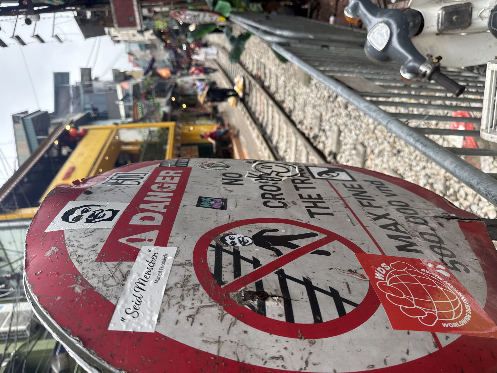 The image shows a close-up, low-angle shot of a weather-beaten, round sign in Hà Nội, Vietnam, with various stickers on it. The sign features a red and white "Danger" warning and a symbol depicting a person crossing out. The sign also states "No Cross The Train" and "Max Fine 500.000 vnd". The sign is located beside active train tracks, a common sight in the city. The backdrop includes glimpses of surrounding buildings and pedestrian activity, typical of Hà Nội's urban landscape. A scooter is partially visible in the background.