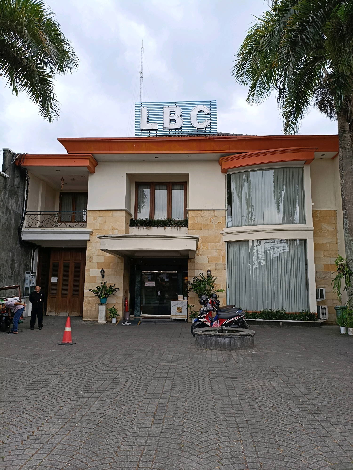 The image shows the exterior of a building with a sign that reads "LBC" at the top. The building has an orange roof trim and stone-colored accents. In front of the building is a paved courtyard with a few potted plants, a security guard in a black uniform standing near a large wooden door, two people attending to some carts, and a red traffic cone. To the right of the entrance, there are two parked motorcycles in front of a circular stone planter. Large windows with sheer curtains cover much of the building's facade. Palm trees frame the scene on the left and right. The sky is overcast. The building is likely located in the city of Depok, Indonesia, as indicated by the "LBC" sign which is associated with a popular Indonesian radio station that has a presence in the area.