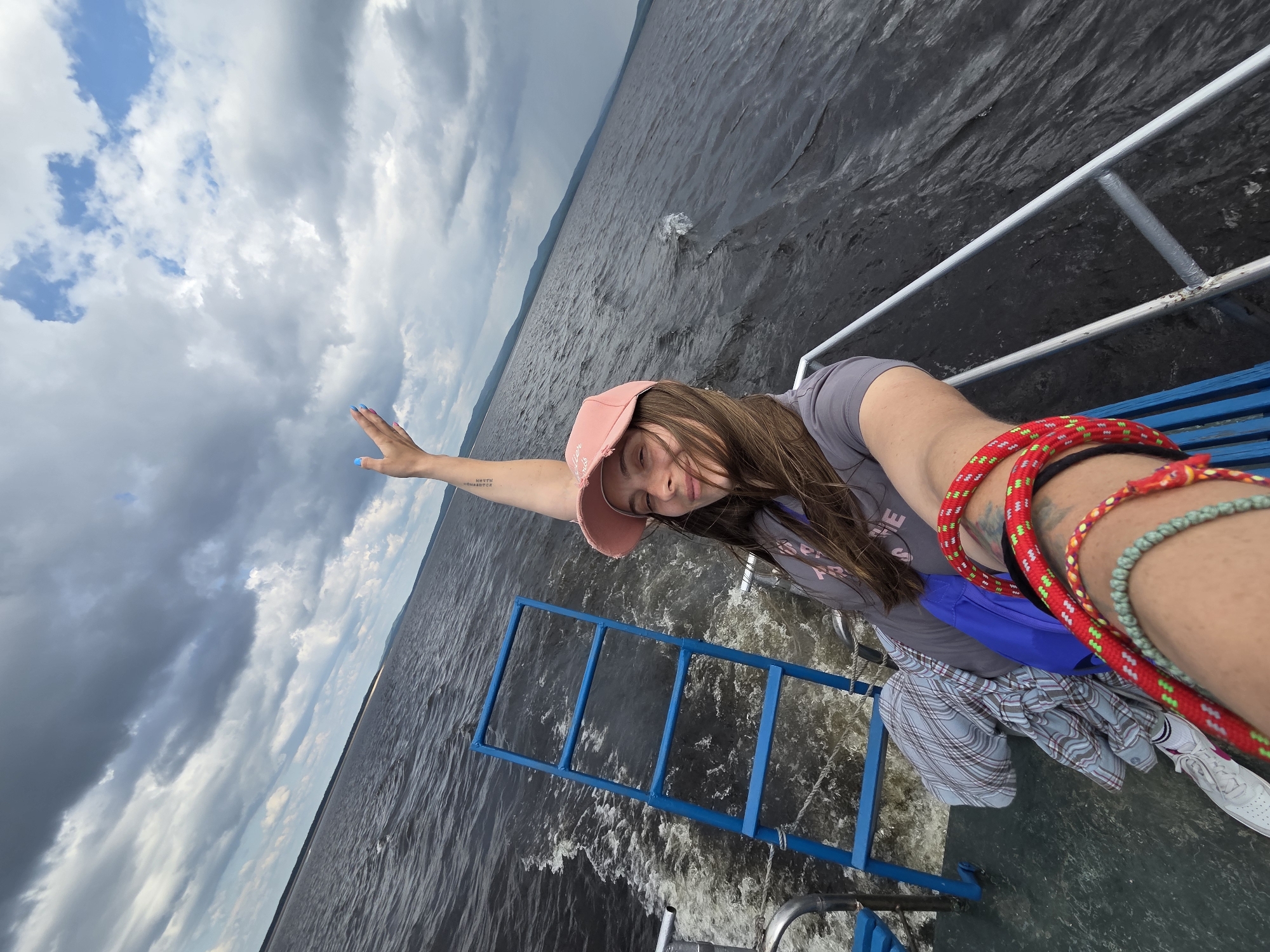 The image shows a young woman on a boat in the city of Talakan, Russia. She is wearing a pink baseball cap and a gray t-shirt. She has her right arm raised, with her hand open and fingers spread. The boat is moving through dark, choppy water, leaving a white wake behind it. The sky is cloudy, with patches of blue visible. In the background, there are hills or mountains. There is a blue ladder on the boat, and a plaid shirt is draped over it. The woman is also wearing several bracelets on her left wrist. The image is taken from a first-person perspective, as if the viewer is on the boat with the woman.