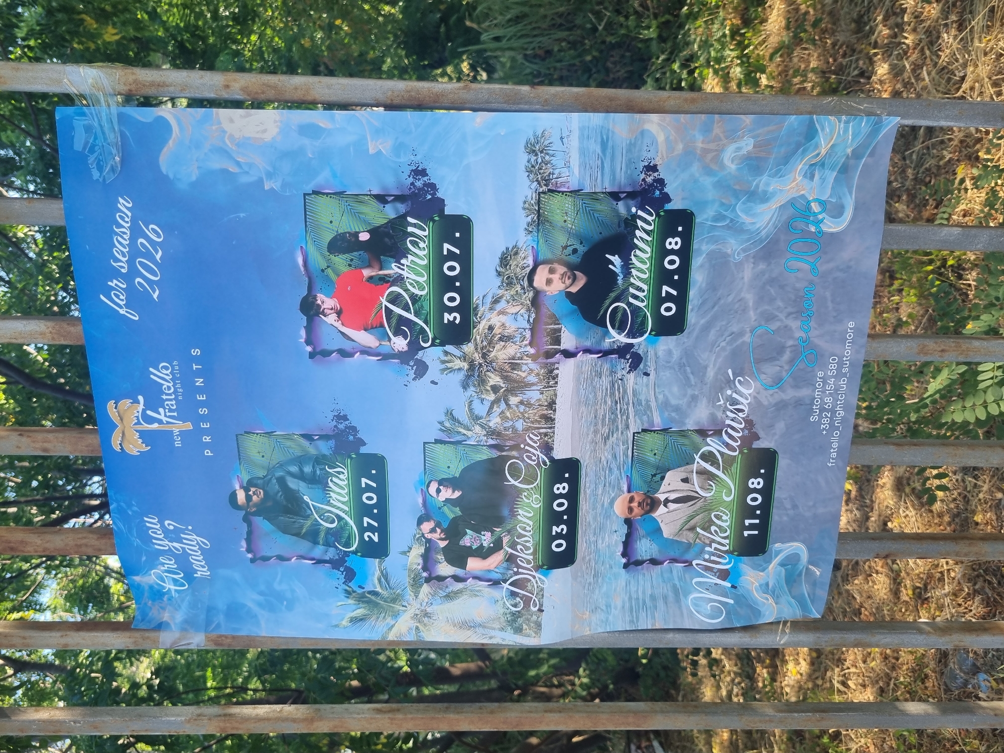A poster for the "Fratello Night Club" is attached to a metal fence outdoors. The poster advertises events for "season 2026" and features images of five performers: Inas on July 27th, Petrov on July 30th, Djekson & Coja on August 3rd, Cunami on August 7th, and Mirko Plavšić on August 11th. The bottom of the poster includes contact information for Sutomore, a phone number, and a website address. The background of the poster depicts a beach scene with palm trees. Trees and vegetation are visible behind the fence, suggesting an outdoor setting.