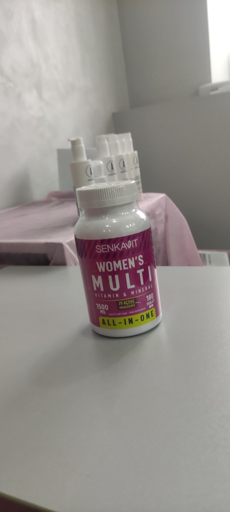 A bottle of Senkavit Women's Multi vitamins stands prominently on a light gray surface. The bottle has a white cap and a pink label with the words "SENKAVIT WOMEN'S MULTI" and information about the contents and ingredients. Behind the vitamin bottle, out of focus, are several white pump bottles on top of a surface covered with a pink cloth. The backdrop is a blurry view of a white wall and a gray wall. The OCR information on the vitamin bottle suggests a Turkish food supplement, potentially indicating that the location is in Russia and is related to the location of the brand. The photograph appears to have been taken in a room with natural light, likely indoors.