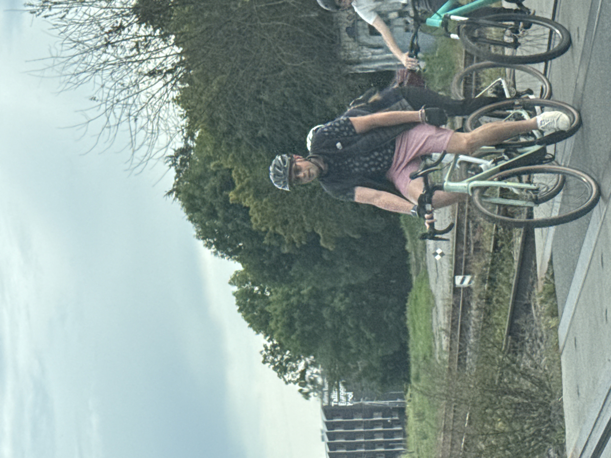 The image captures a scene with three cyclists on a paved surface, possibly a crossing, with railroad tracks visible in the background. The sky is overcast and light blue with scattered clouds. To the left, a large, dark building with a repeating pattern of vertical lines and horizontal accents is partially visible, with some white text that appears to read "Tallinn Europort." Dense green trees line the mid-ground, partially obscuring the building and the area behind the railroad tracks.
In the foreground, the primary subject is a man wearing a dark helmet, sunglasses, and a black vest over a patterned shirt. He is also wearing pink shorts and white athletic shoes, and is riding a light green bicycle. His left hand is on the handlebars, and his legs are pedaling. Behind him, another cyclist, mostly obscured, is riding a bicycle, wearing dark clothing. To the right, a third cyclist is visible, wearing a white t-shirt and black cycling shorts, riding a turquoise bicycle. Their right hand is on the handlebars, and they are also in motion. The railroad tracks have several wooden ties and metal rails, with some green grass and brush growing around them. A white and black marker is visible on the tracks in the mid-ground.