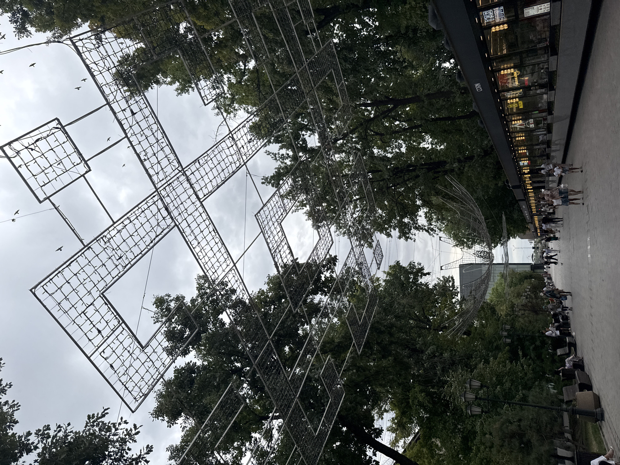 The image depicts a street scene in Shymkent, Kazakhstan. Overhead, a grid-like structure of metal frames and lights is suspended across the sky, with some elements resembling geometric shapes. Numerous small birds are visible in flight against the overcast sky. Tall, leafy trees line both sides of the street. On the right side of the street, there is a long, modern building with a dark facade and a row of illuminated storefronts displaying signage and reflections. People are walking along the paved pedestrian walkway. On the left side of the street, benches and lampposts are situated among the trees. The overall atmosphere suggests an urban public space on a cloudy day.