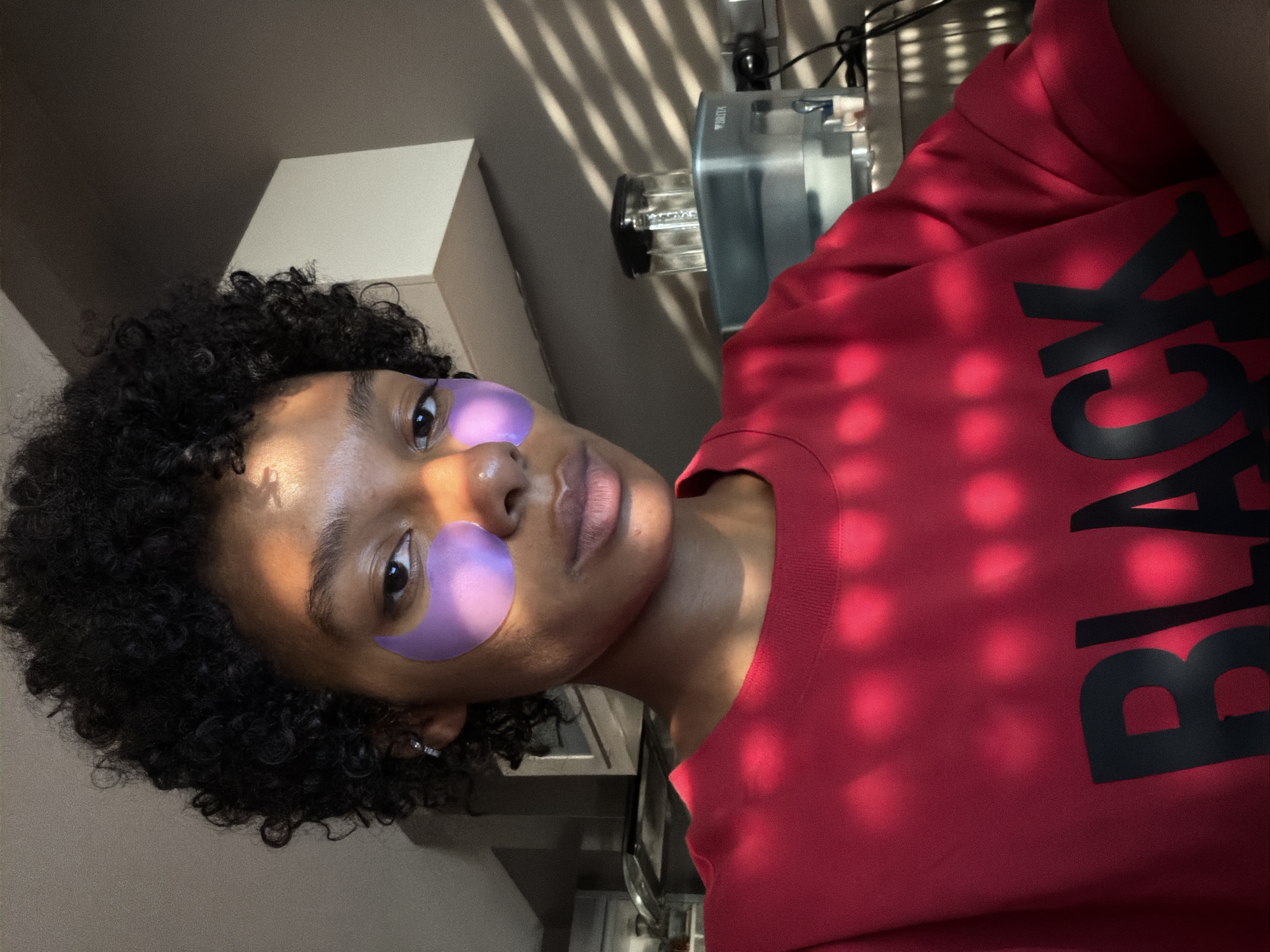 A young Black woman with short, curly dark hair is shown from the chest up, looking directly at the camera. She is wearing a bright red t-shirt with the word "BLACK" printed in large black letters across the front. She has purple under-eye patches on her face. Sunlight streams through a window with blinds, casting diagonal shadows across her face and t-shirt. In the background, a Brita water filter is visible on a kitchen counter. The scene appears to be indoors, likely in a kitchen, in Champs-sur-Marne, France.