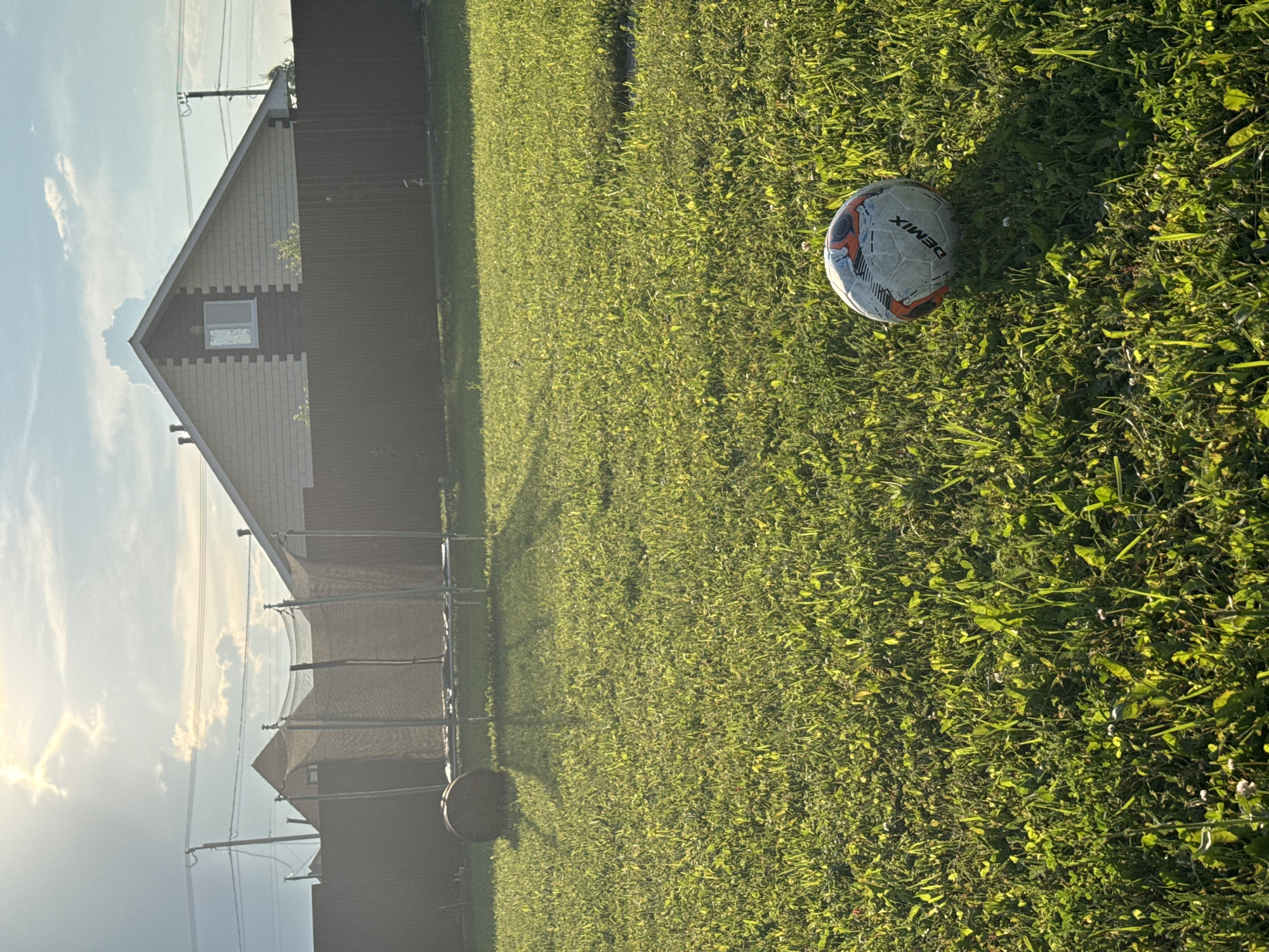 The image depicts a sunny backyard scene in Zav'yalovo, Russia, with a well-maintained lawn. In the foreground, a soccer ball with the brand "DEMIX" is resting on the grass. In the midground, there is a trampoline with a safety net. Behind the trampoline, a dark brown fence encloses the yard. A portion of a residential building, featuring a light-colored facade with darker trim and a single window in the gable, is visible above the fence. The sky is bright with scattered clouds, and the sun is shining, casting long shadows across the lawn. A utility pole with power lines is also present in the background.