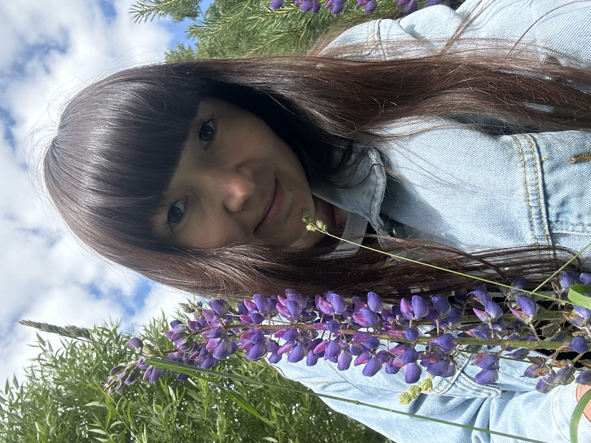 The image shows a close-up of a woman with dark brown hair and bangs, wearing a light blue denim jacket. She is outdoors, surrounded by lush green foliage and purple lupine flowers. The sky is visible in the background with scattered white clouds. The lighting suggests it is daytime. The woman is looking towards the camera with a slight smile. This scene appears to be in a natural setting, possibly a park or a field, consistent with the natural beauty found in areas like the banks of the Kama River in Chaikovsky, Russia, which are known for their scenic landscapes and recreational spaces.