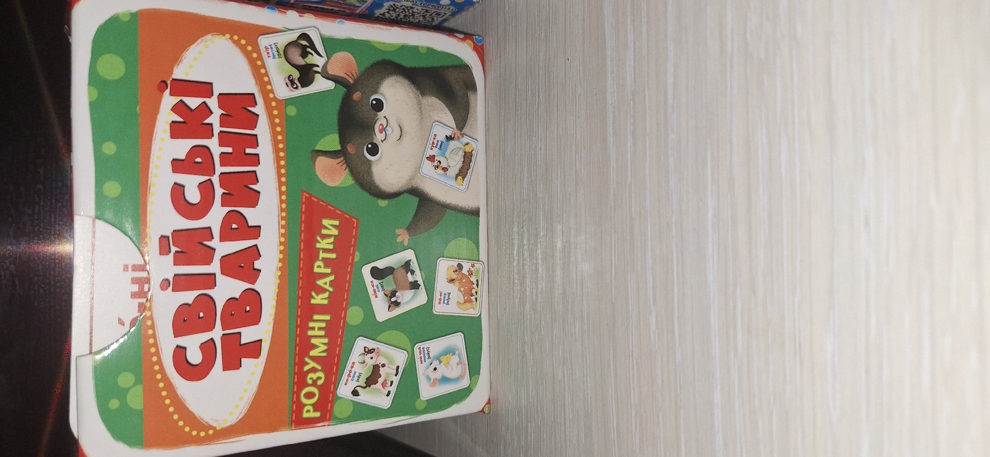 The image shows a close-up of a board game box featuring cartoon animals and Ukrainian text. The text on the box reads "СВІЙСЬКІ ТВАРИНИ" (Domestic Animals) and "РОЗУМНІ КАРТКИ" (Smart Cards). Several smaller cards with images of animals like a cow, cat, mouse, horse, chicken, and ferret are scattered around a central hamster character. The box is primarily colored orange and green with yellow polka dots. The bottom portion of the image reveals a light-colored wooden surface, possibly a table. There is no indication of the city of Bila Tserkva, Ukraine, in the image.