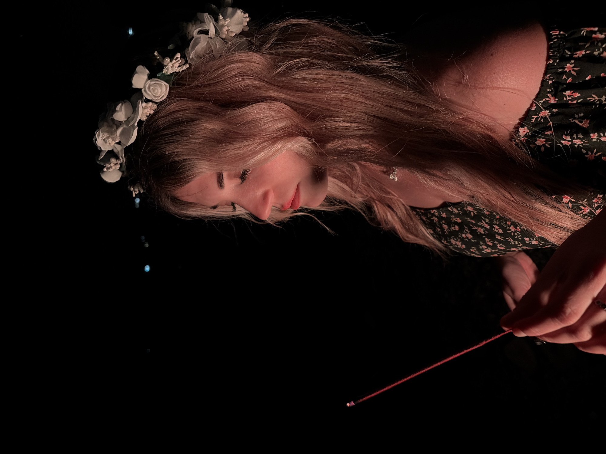 A young woman with long blonde hair adorned with a white floral crown looks down at a thin, red sparkler she holds in her hands. She is wearing a dark, floral print dress. The background is entirely black. There are no visible elements in the picture to indicate the location. Therefore, the location is unknown, and Kamenovo, Montenegro, cannot be confirmed.