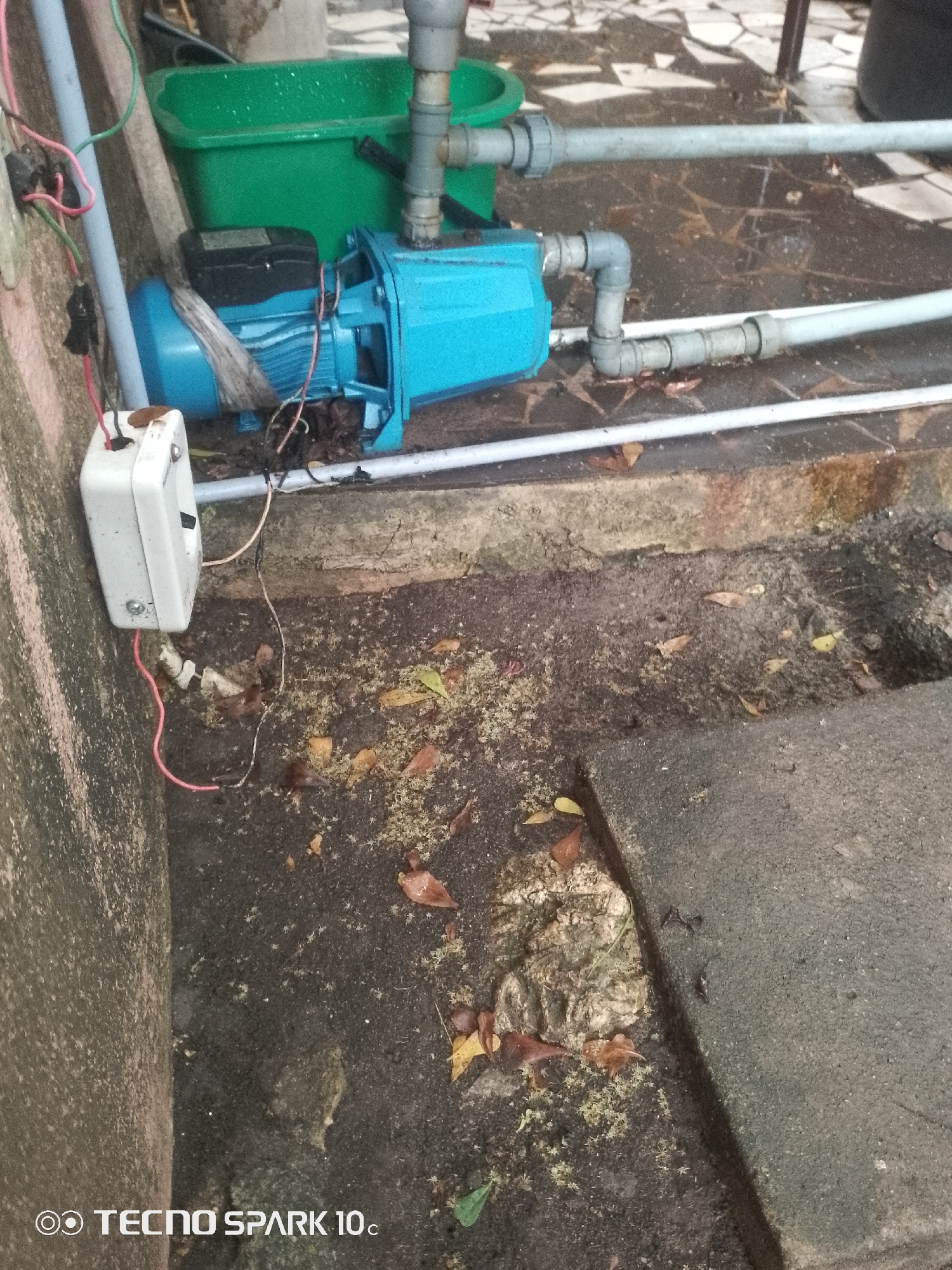 The image shows an outdoor scene in Lekki, Nigeria. In the foreground, a blue water pump is situated next to a green plastic tub. Electrical wires connect the pump to a white switch box mounted on a textured wall. The ground is a mix of concrete, dirt, and scattered dry leaves. The overall environment appears to be a utilitarian or service area, possibly in a residential or commercial setting within Lekki.