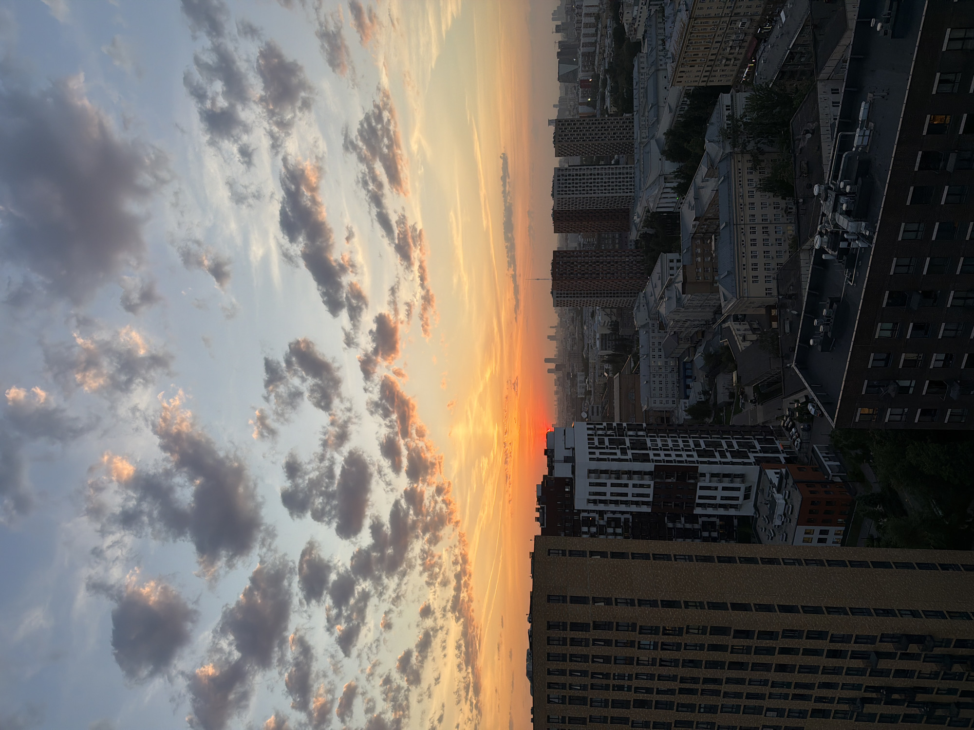 The image depicts a sunset over an urban landscape, likely a city. The sky is filled with clouds tinged with orange and pink hues, indicating the setting sun. The buildings below vary in height and architecture, with some modern high-rises and older, shorter structures. Details of people and specific actions are not discernible due to the distance and focus on the overall scene. The environment is urban, with a skyline dominated by buildings. Location is Unknown, Unknown.