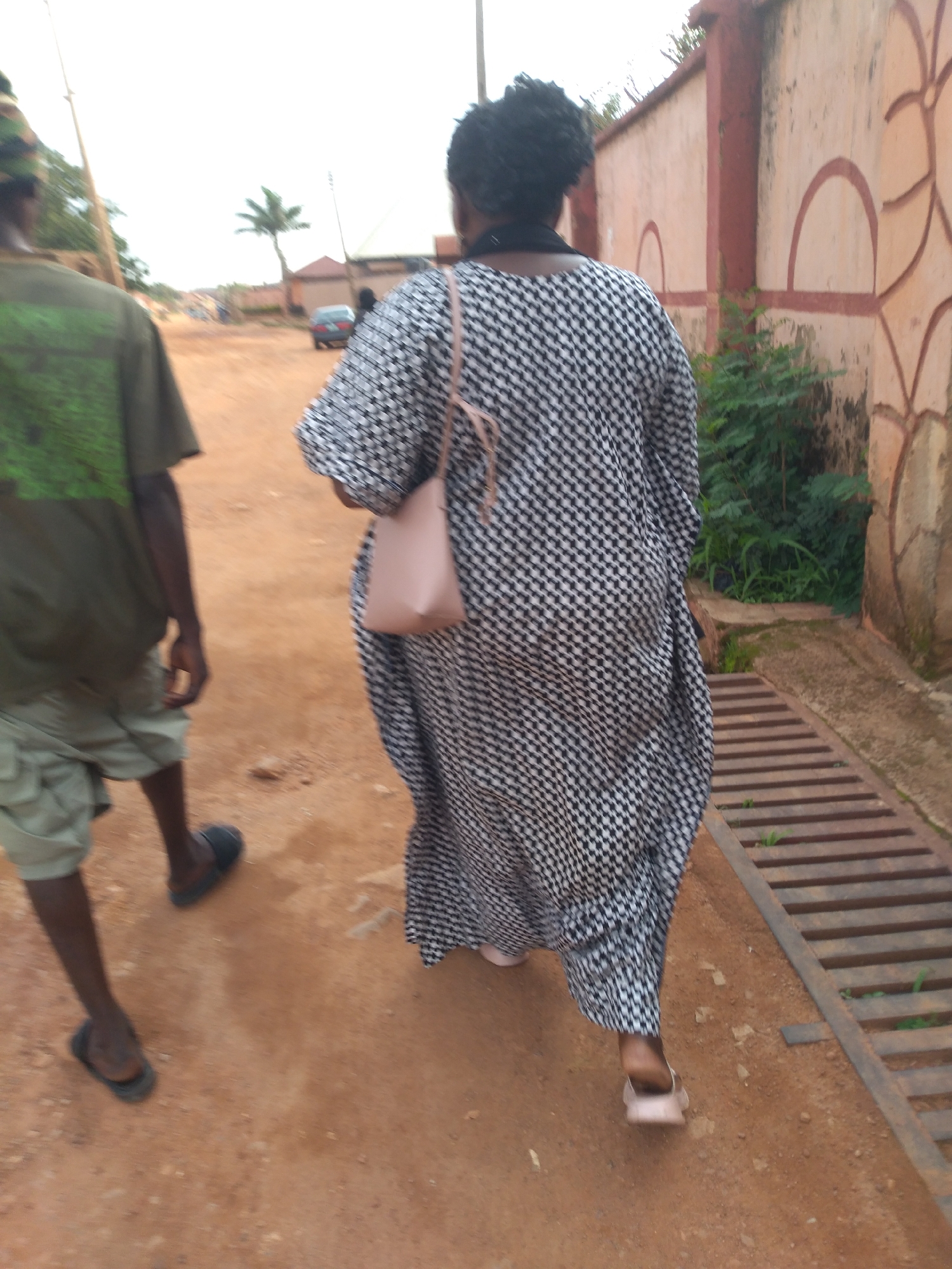 In Barakin Akawo, Nigeria, two individuals are walking on a dirt road adjacent to a textured concrete wall. The person in the foreground, viewed from the rear, wears a black and white checkered maxi dress and carries a light pink handbag. Their feet are bare, with a hint of a light-colored shoe. To their left, another individual, also viewed from the rear, is dressed in a green t-shirt and light-colored shorts, wearing dark sandals. Their right leg is slightly ahead of their left. The environment appears to be a street or pathway, with a metal grate running along the right side of the image. Vegetation is visible behind the wall and near the grate. In the background, a car is parked further down the dirt road, and a palm tree stands against a hazy sky. The ground is composed of reddish-brown soil with scattered pebbles.